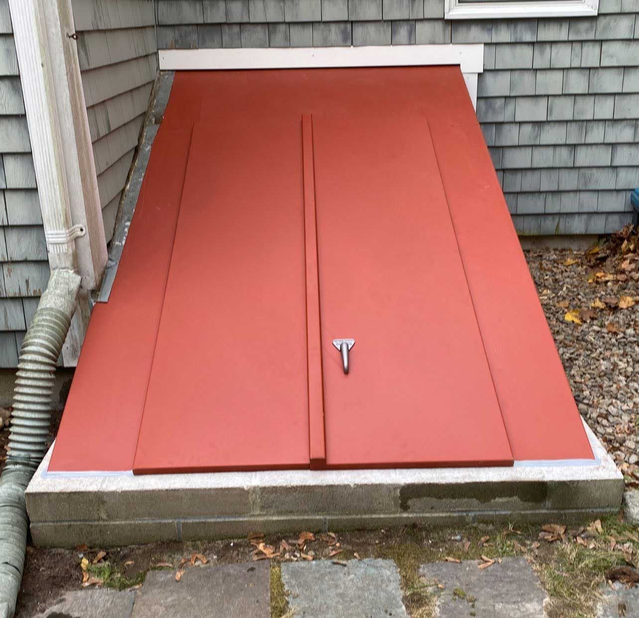 Red cellar doors on a concrete base, next to a house with gray siding. A downspout is on the left.