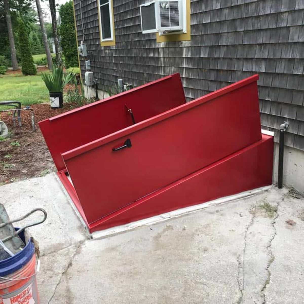 Red metal basement hatch doors, open on concrete beside a gray house.