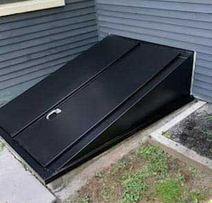 Black metal basement hatch angled against gray siding, surrounded by grass and concrete.