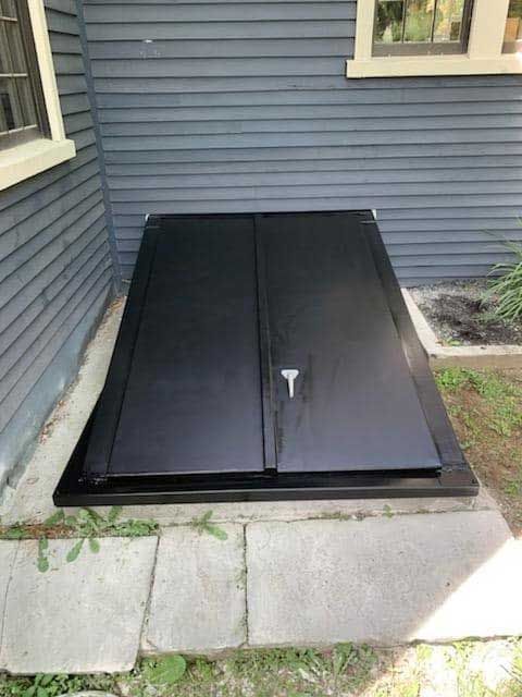 Black cellar door set in concrete near a blue house wall with a window.