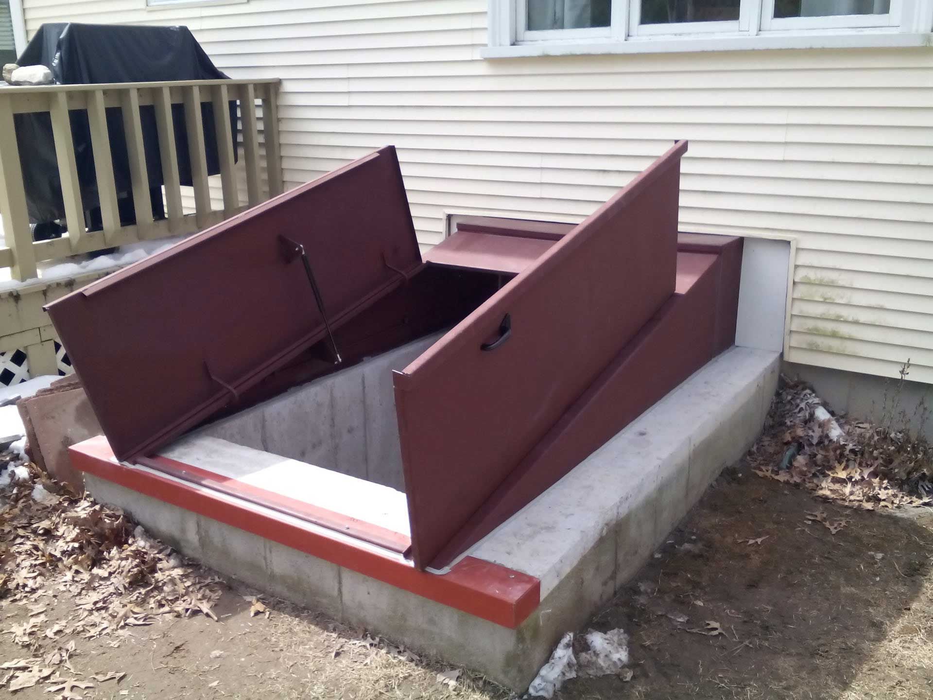 Burgundy metal basement door open, built into a concrete foundation next to a house.