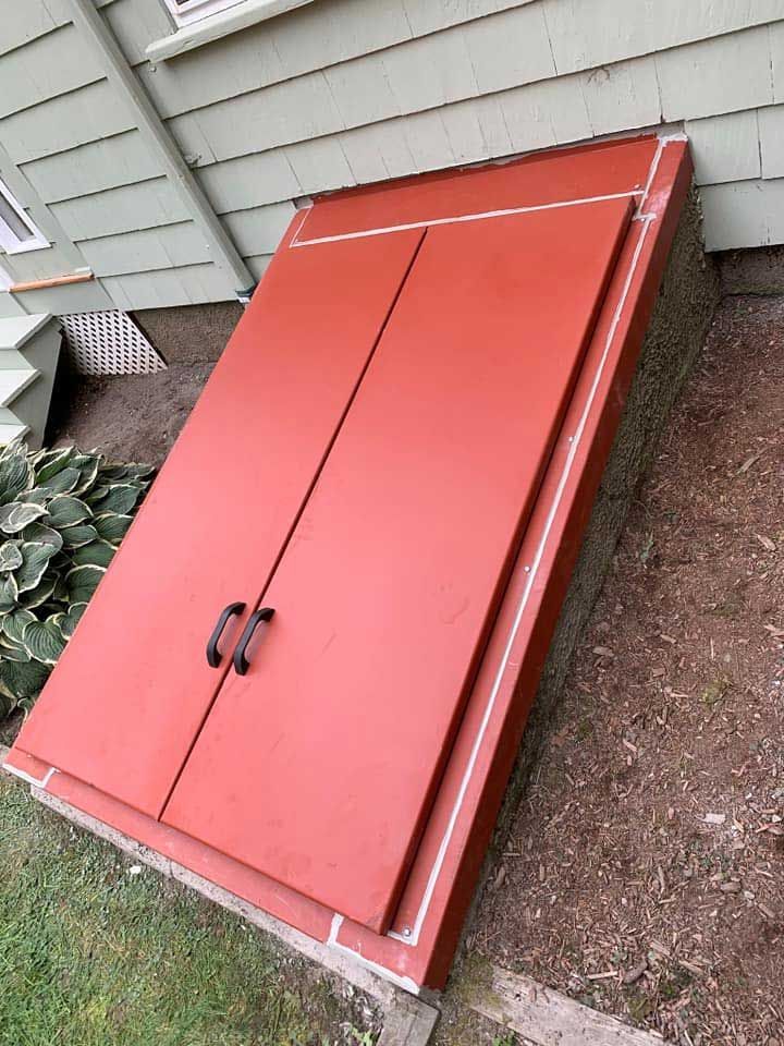 Red cellar doors against a light green house, surrounded by grass and dirt.