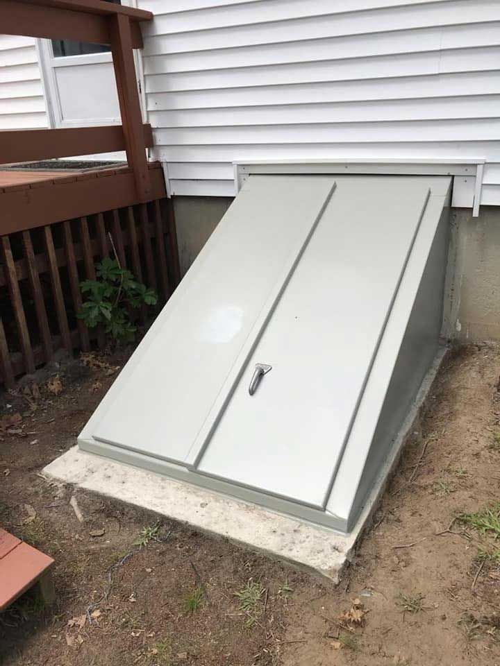 Gray cellar door set in concrete, against white siding, beside a deck.