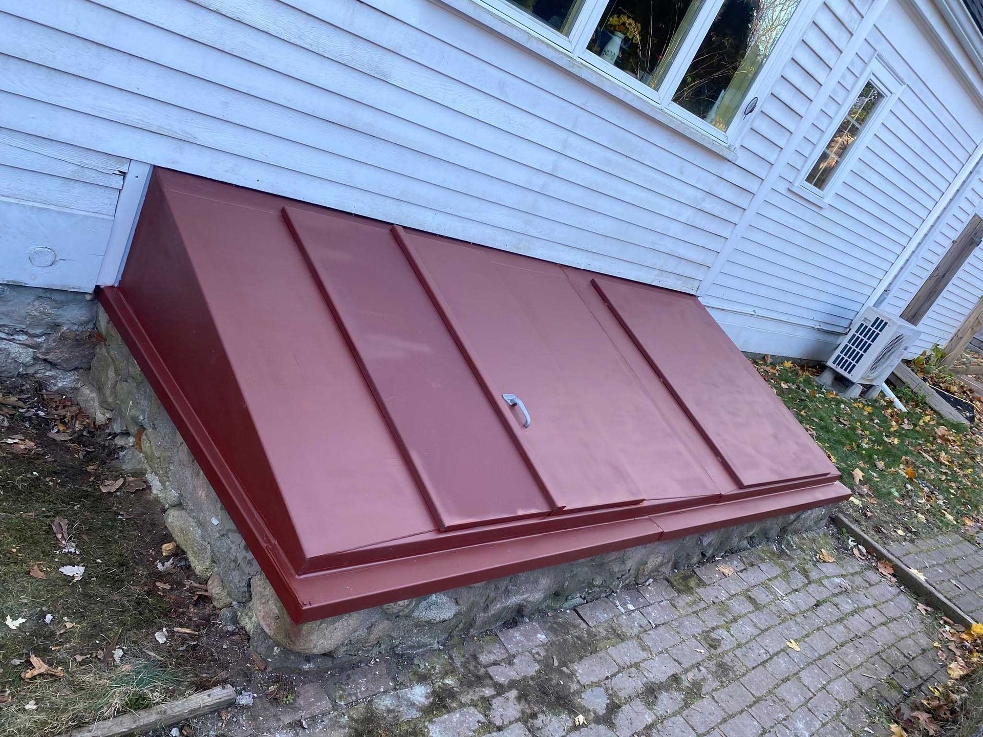 Red metal basement door on a stone foundation against a white house with a window and a small air conditioner.