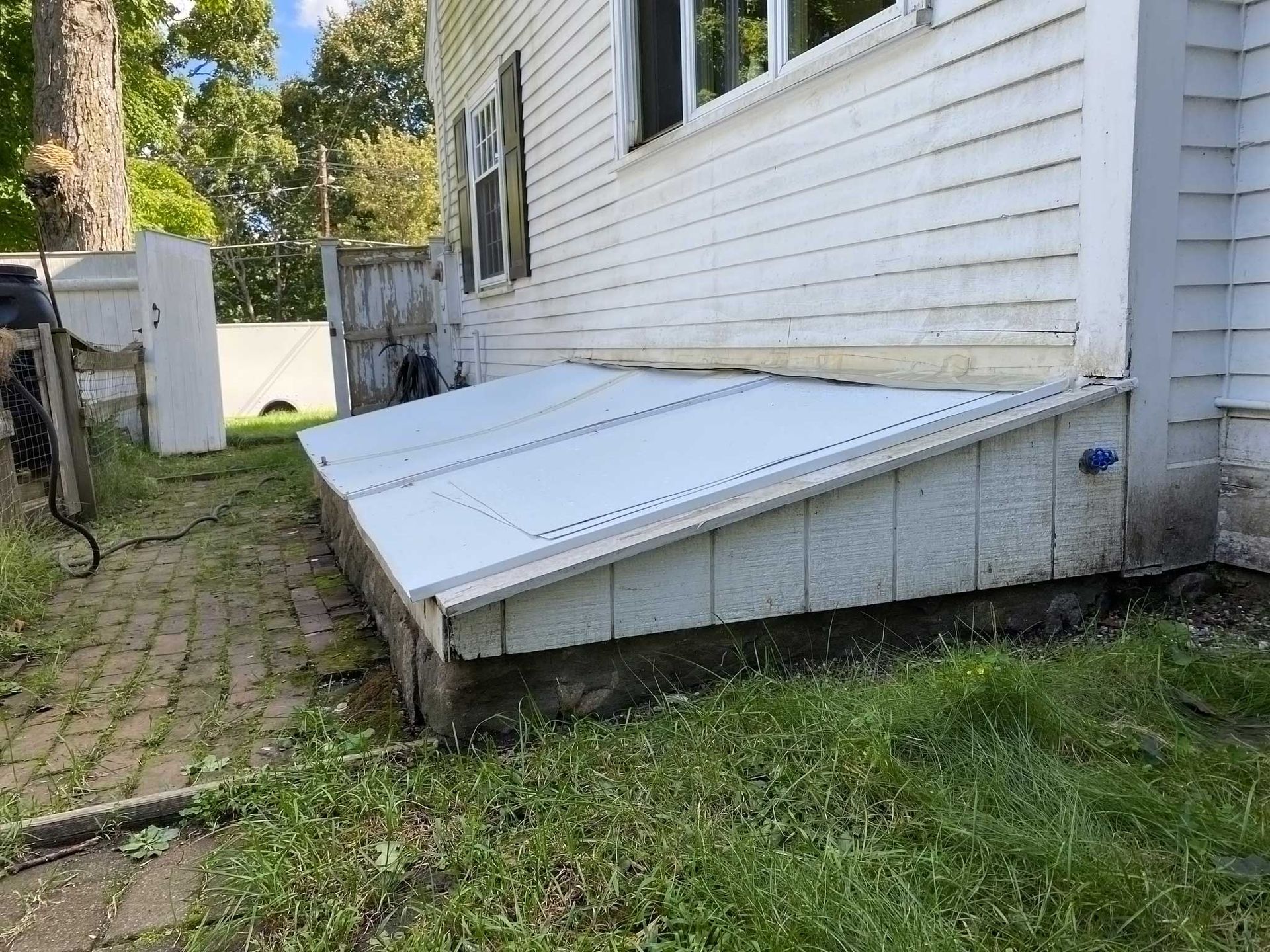 White-sided house with a white metal cellar door. A brick path and green grass are in the foreground.