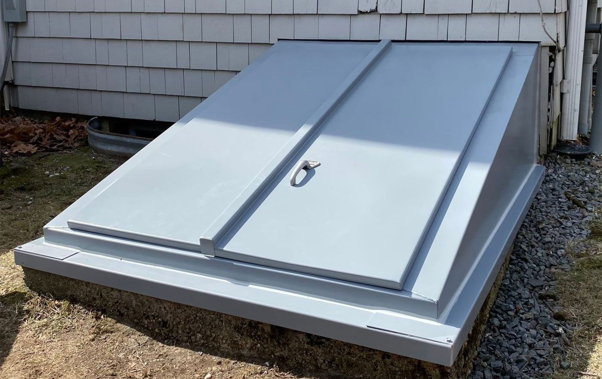 Gray, sloped basement entry doors on a concrete base, set against a white-sided building and a gravel yard.