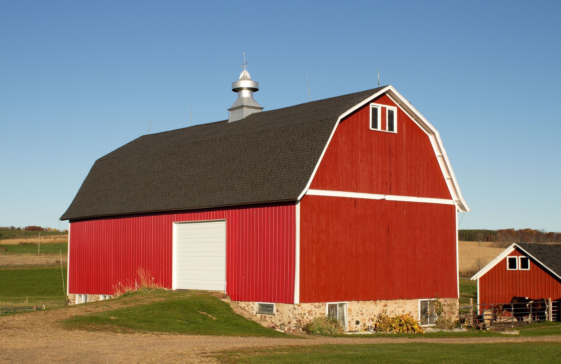 A red barn with a gambrel roof and a white cupola sits in a rural field under a clear blue sky.