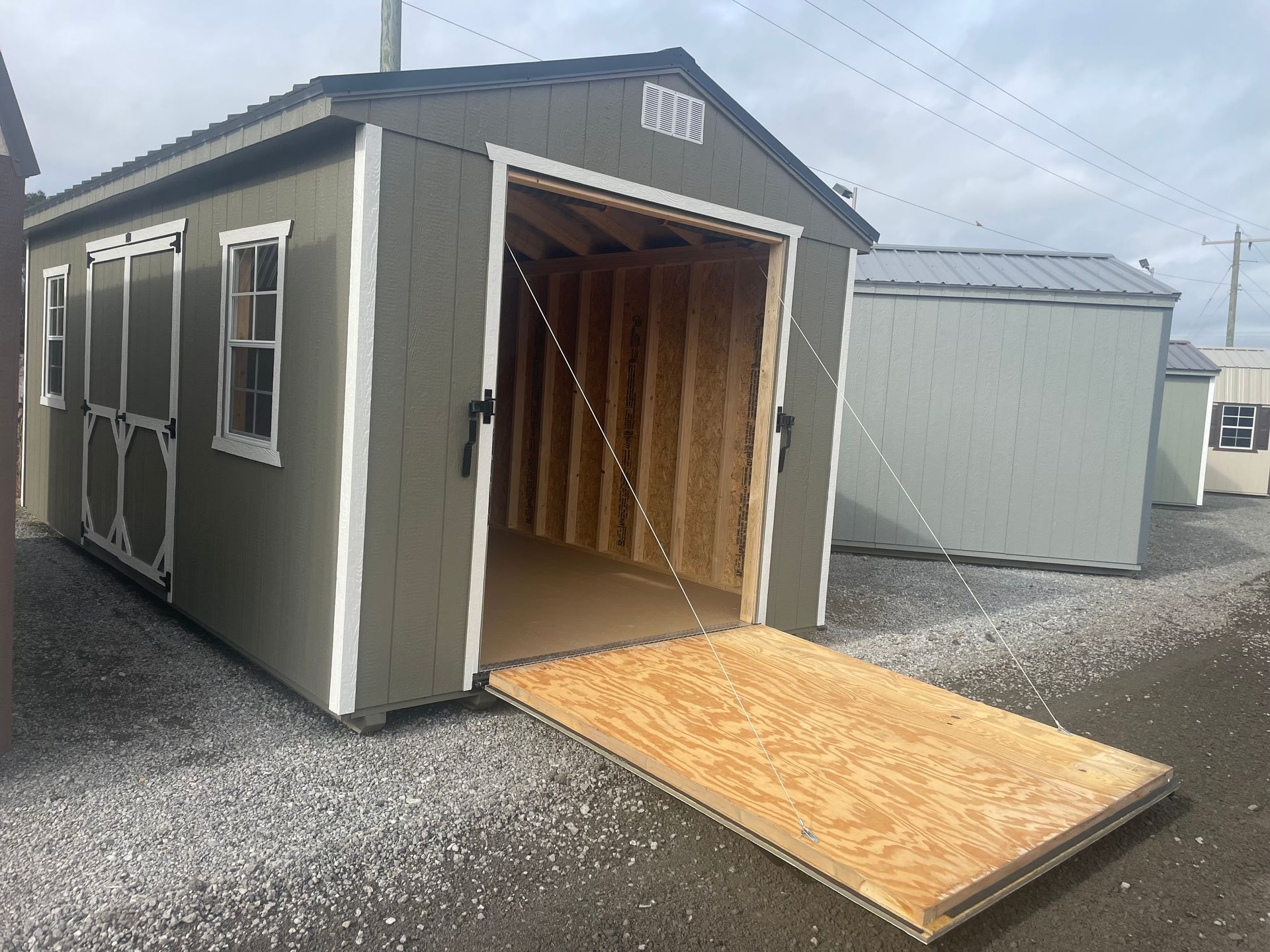 Green storage shed with a wooden ramp lowered at the entrance, situated in an outdoor lot with gravel ground.