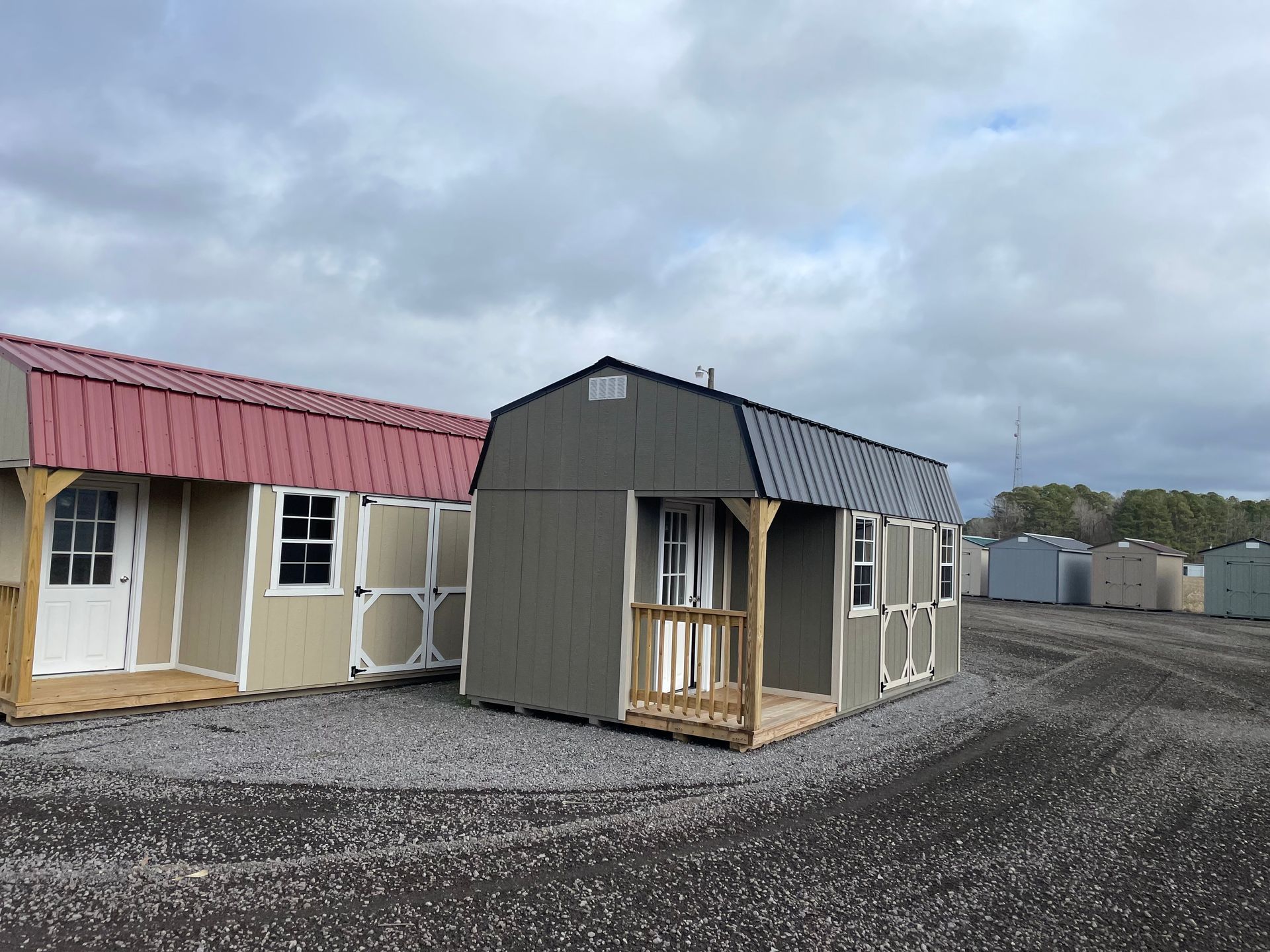 Two portable storage sheds with porches, one with a red metal roof and the other grey, sit on a gravel lot under a cloudy sky.
