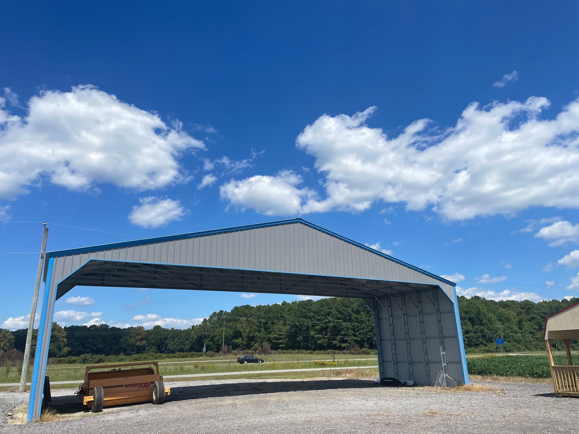 A metal carport stands on a gravel lot under a bright blue, cloudy sky, with a wooden trailer tucked under its frame.