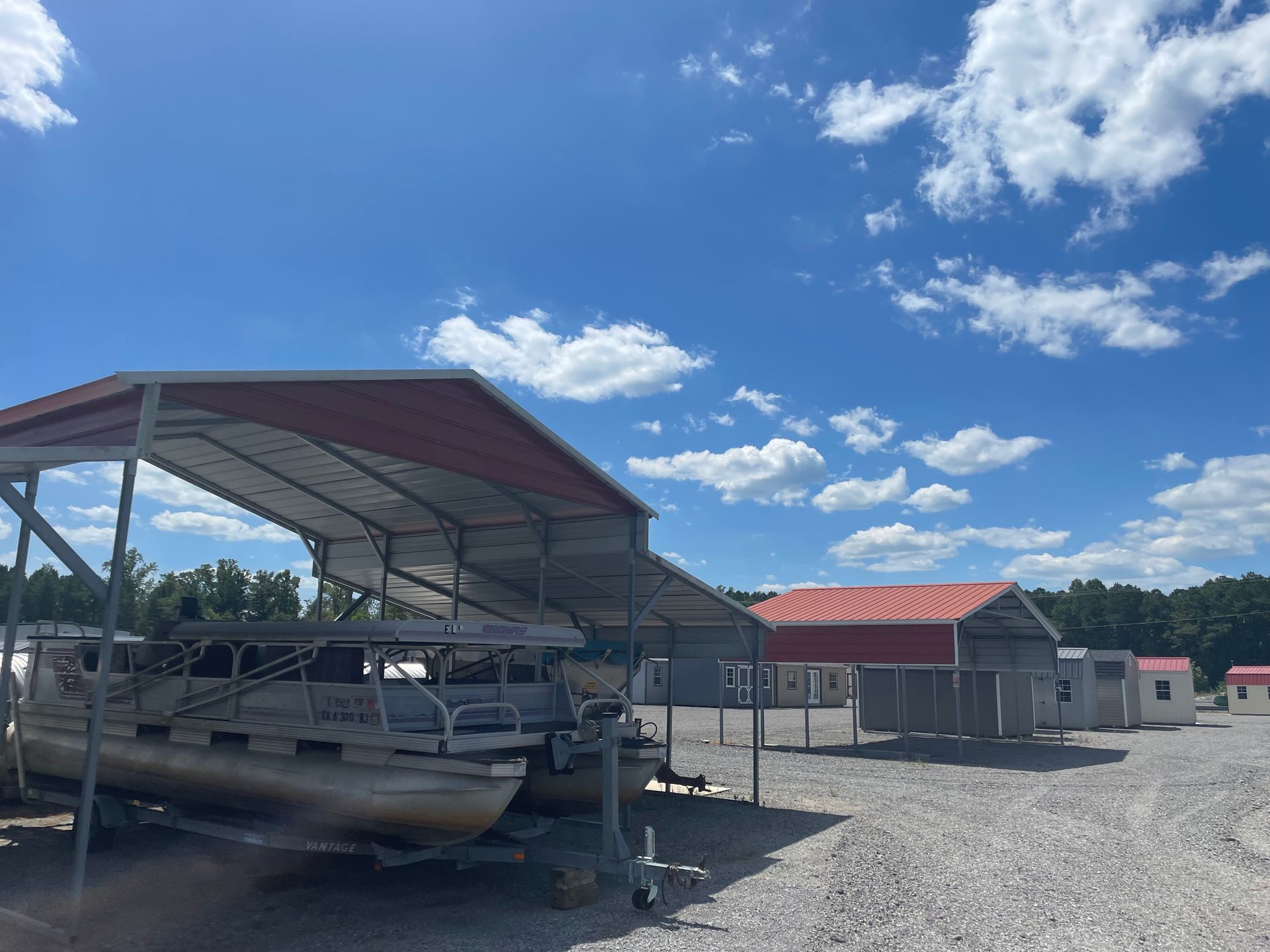 A pontoon boat parked under a metal carport in a gravel lot with additional storage sheds under a sunny, cloudy sky.