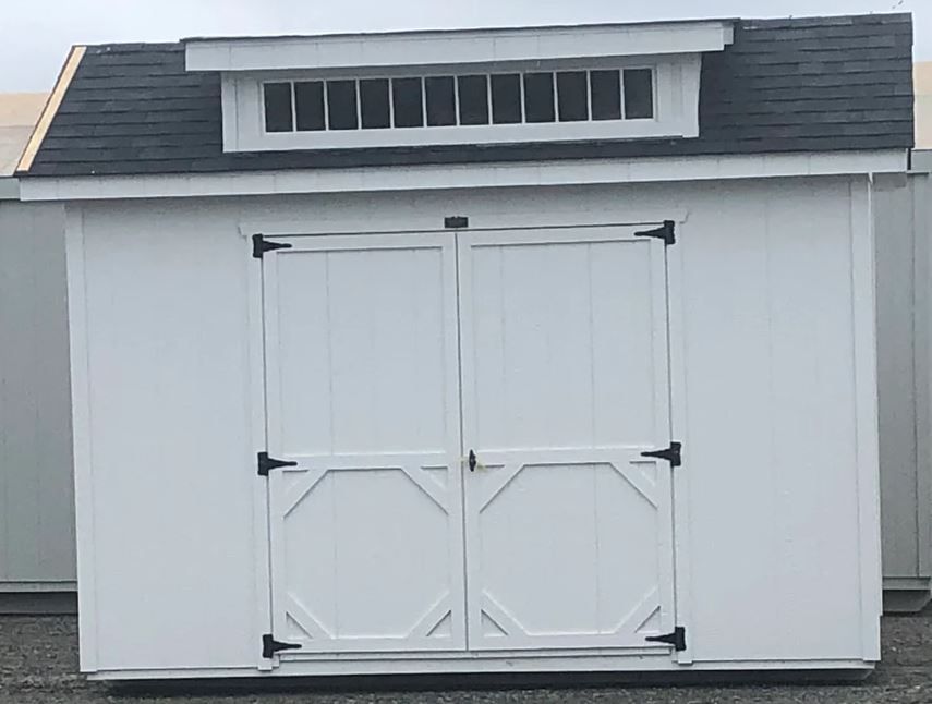 A white shed with black roof shingles and a row of transom windows above double doors.