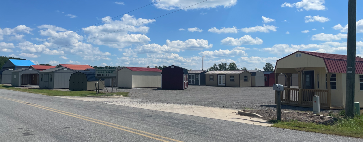 A gravel lot displaying various pre-built storage sheds and tiny homes under a blue sky with scattered clouds.