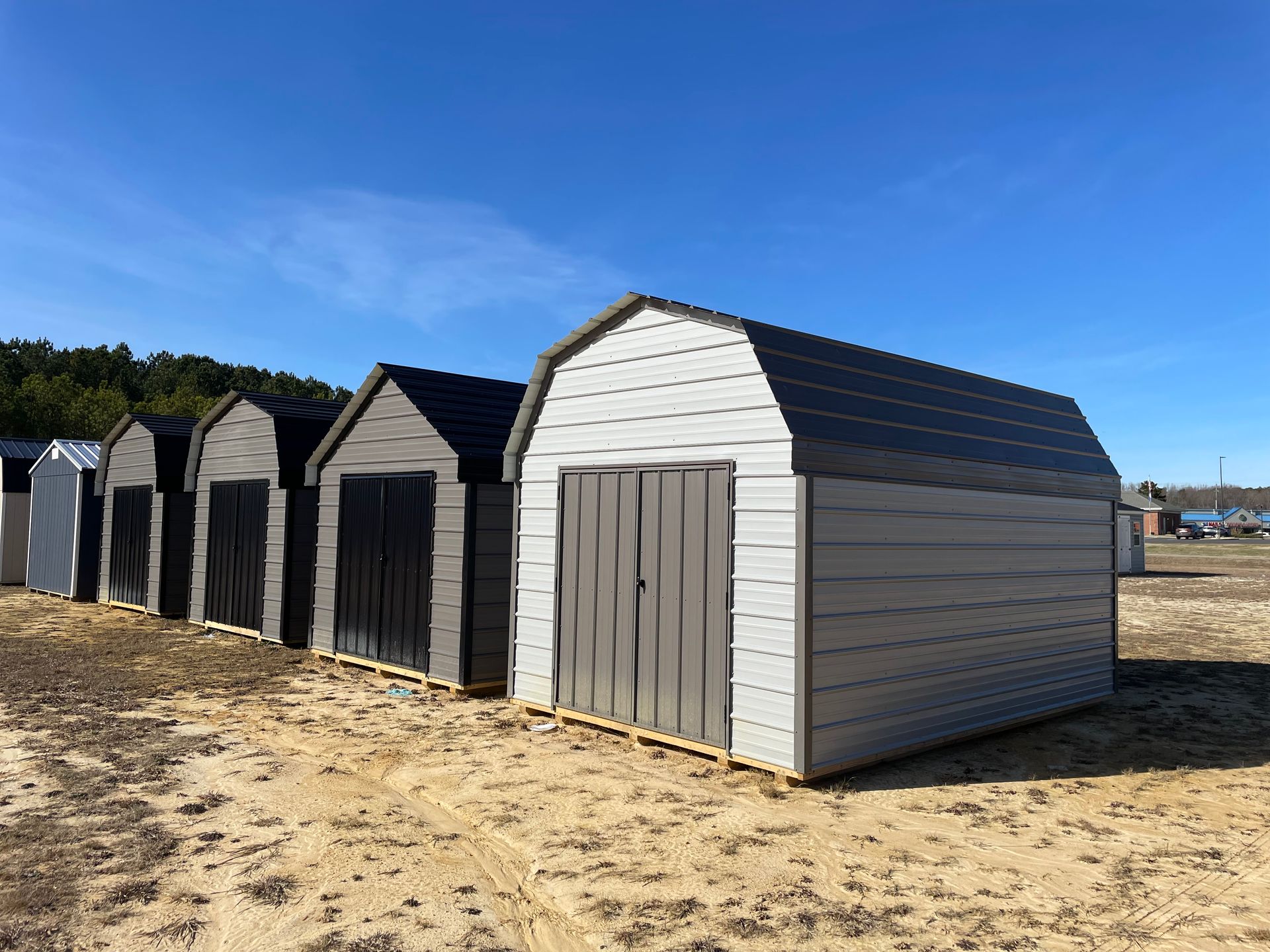 A row of new, light gray and dark gray storage sheds sitting in a dry, sandy field under a clear blue sky.