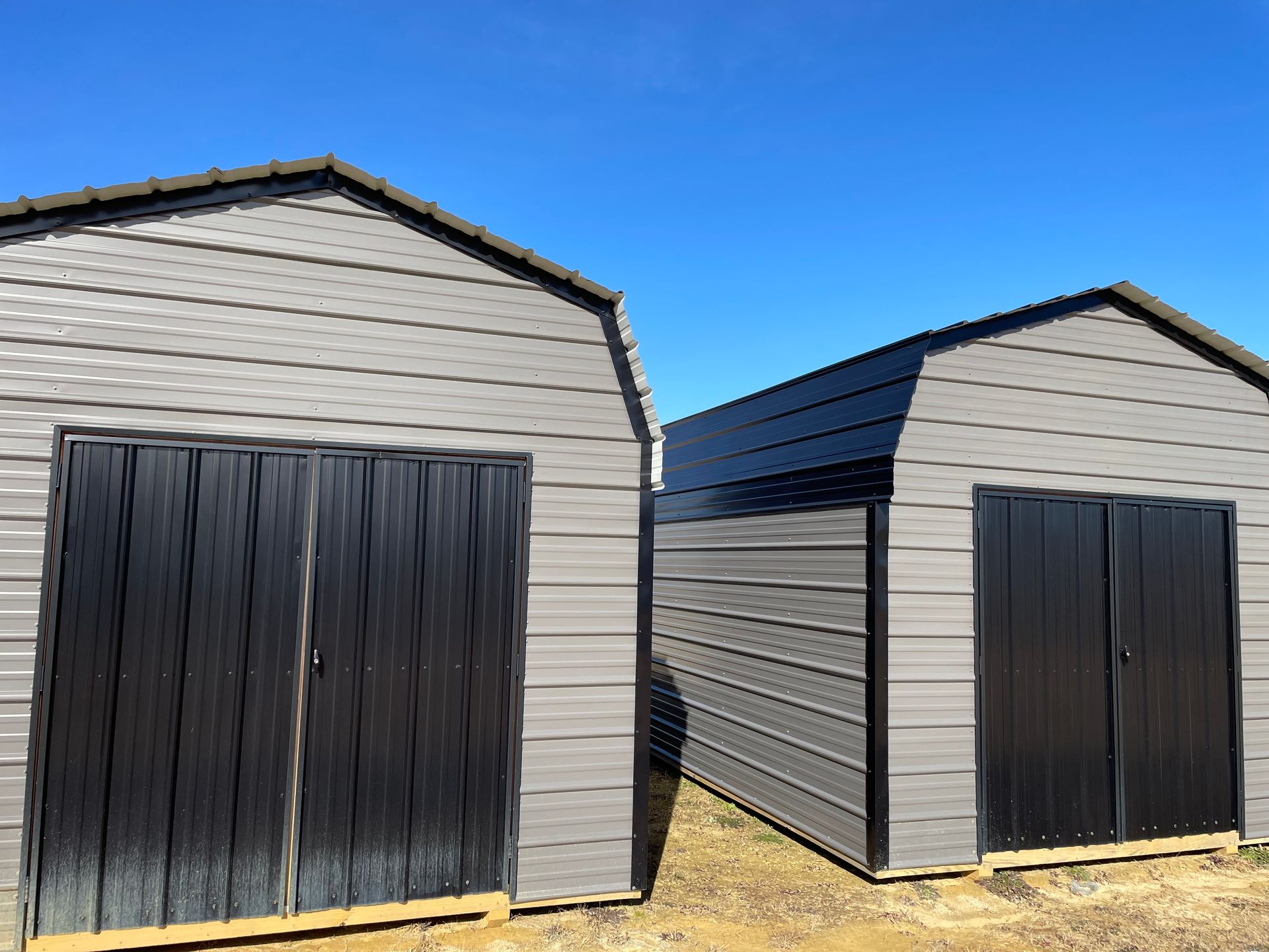 Two gray metal storage sheds with black double doors stand side-by-side against a clear blue sky.
