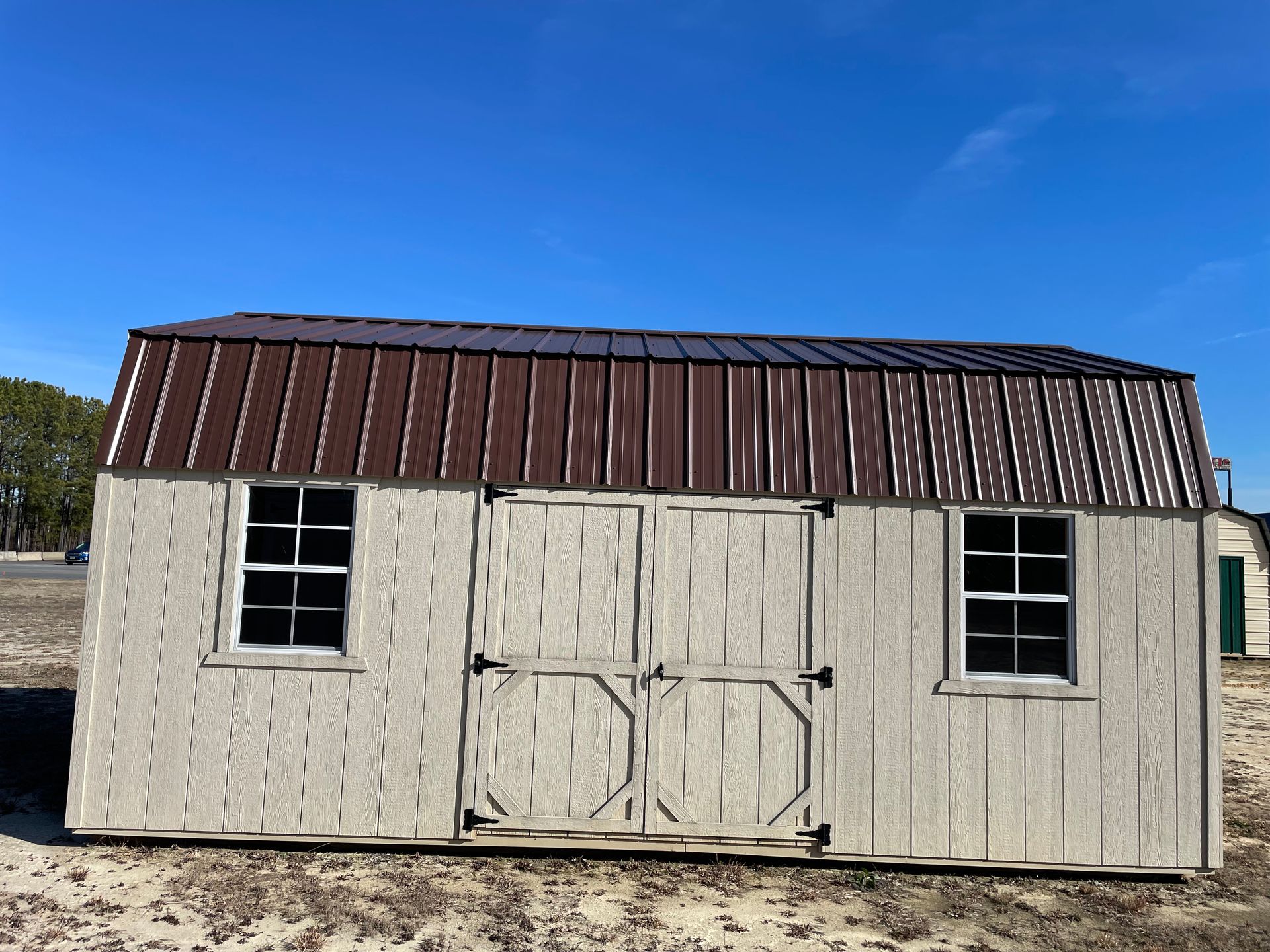 A light-colored shed with a dark metal gambrel roof and double doors, situated in a sandy outdoor lot under a clear sky.