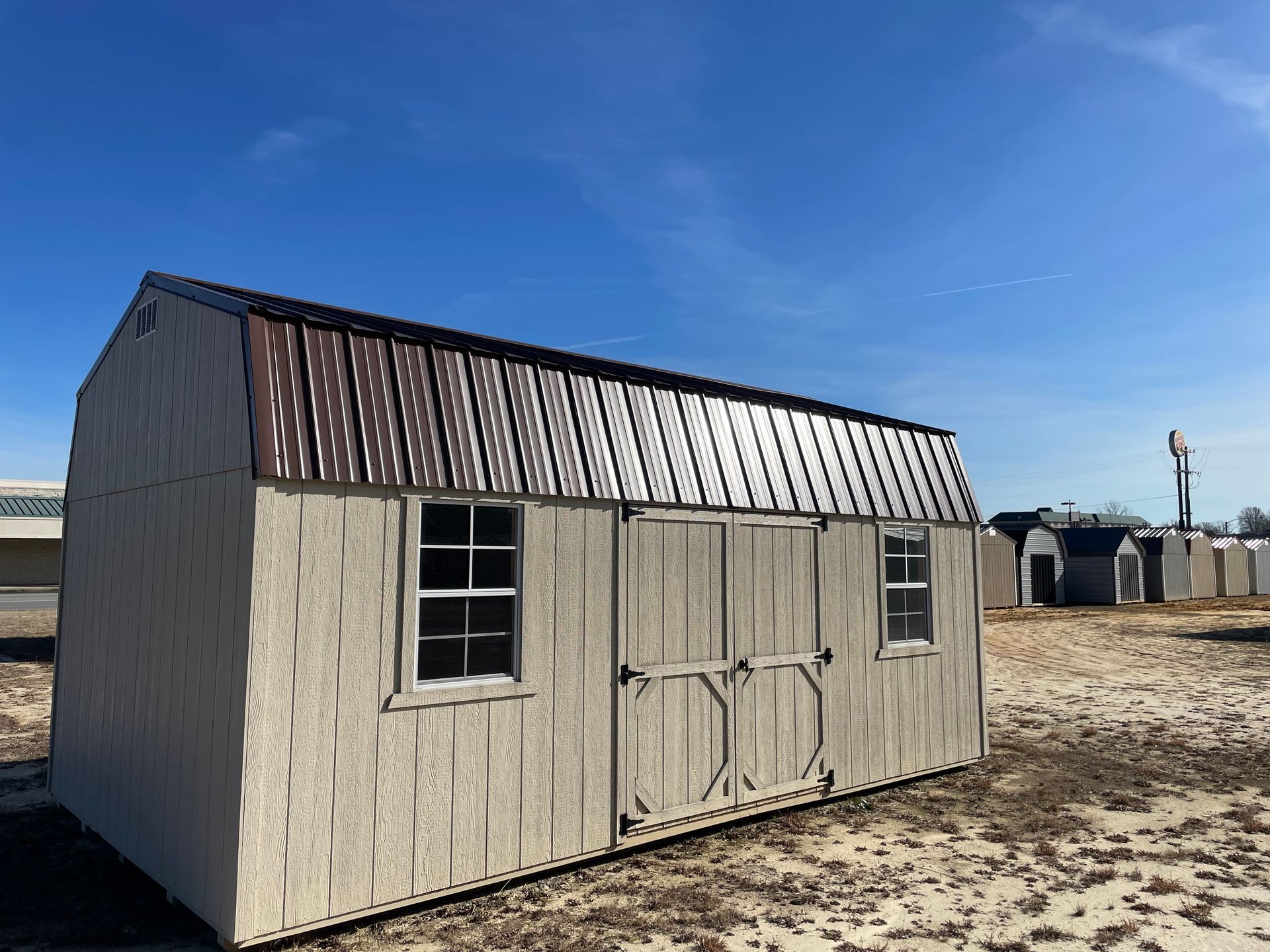 A beige wooden shed with a brown metal gambrel roof and double doors, sitting on a gravel lot under a clear blue sky.