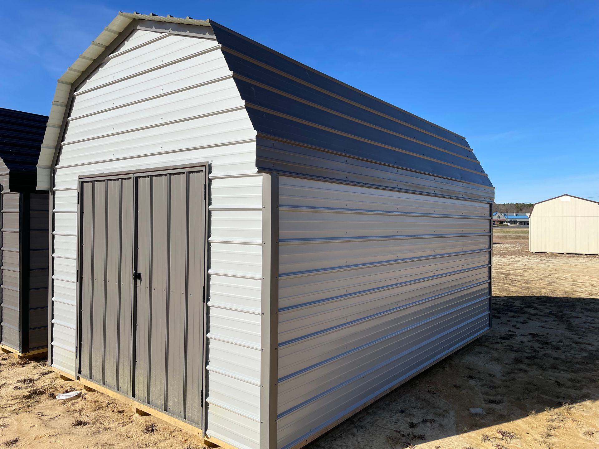 A metal barn-style storage shed with light grey siding and a dark grey roof and double doors in a sunny, open field.