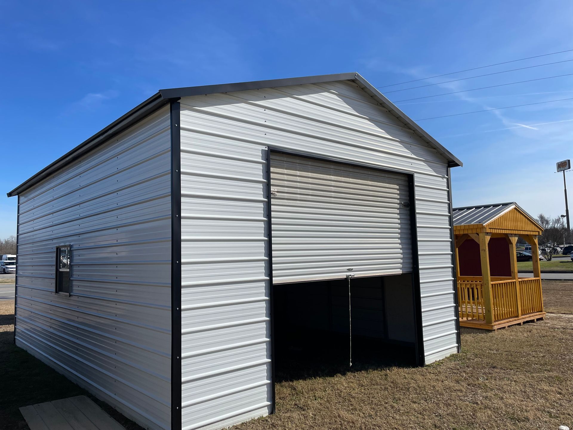 A white metal storage shed with a roll-up garage door, located in an outdoor lot next to a yellow wooden structure.