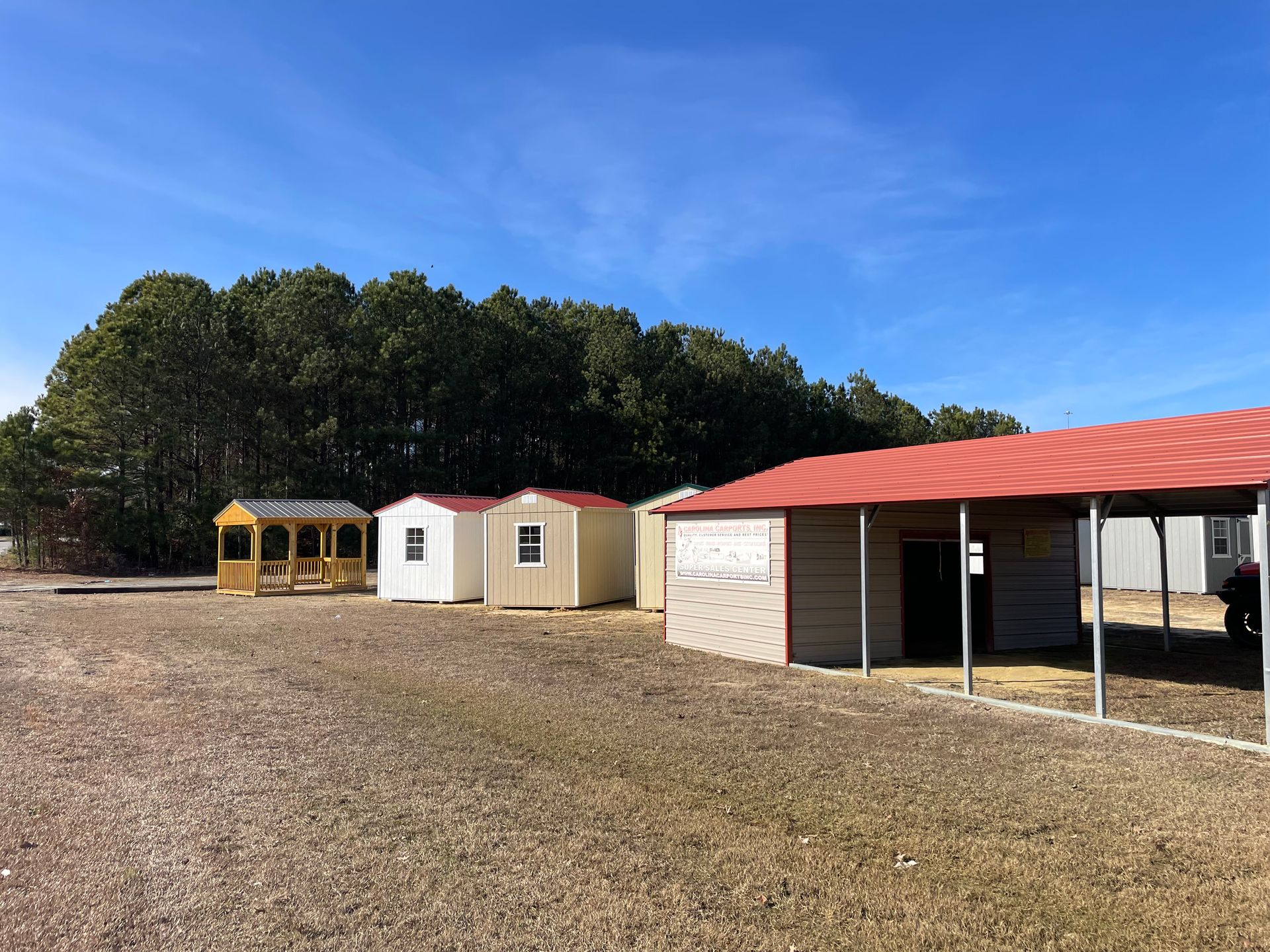 A row of small storage sheds and a larger metal-roofed shelter on a gravel lot in front of a line of trees.