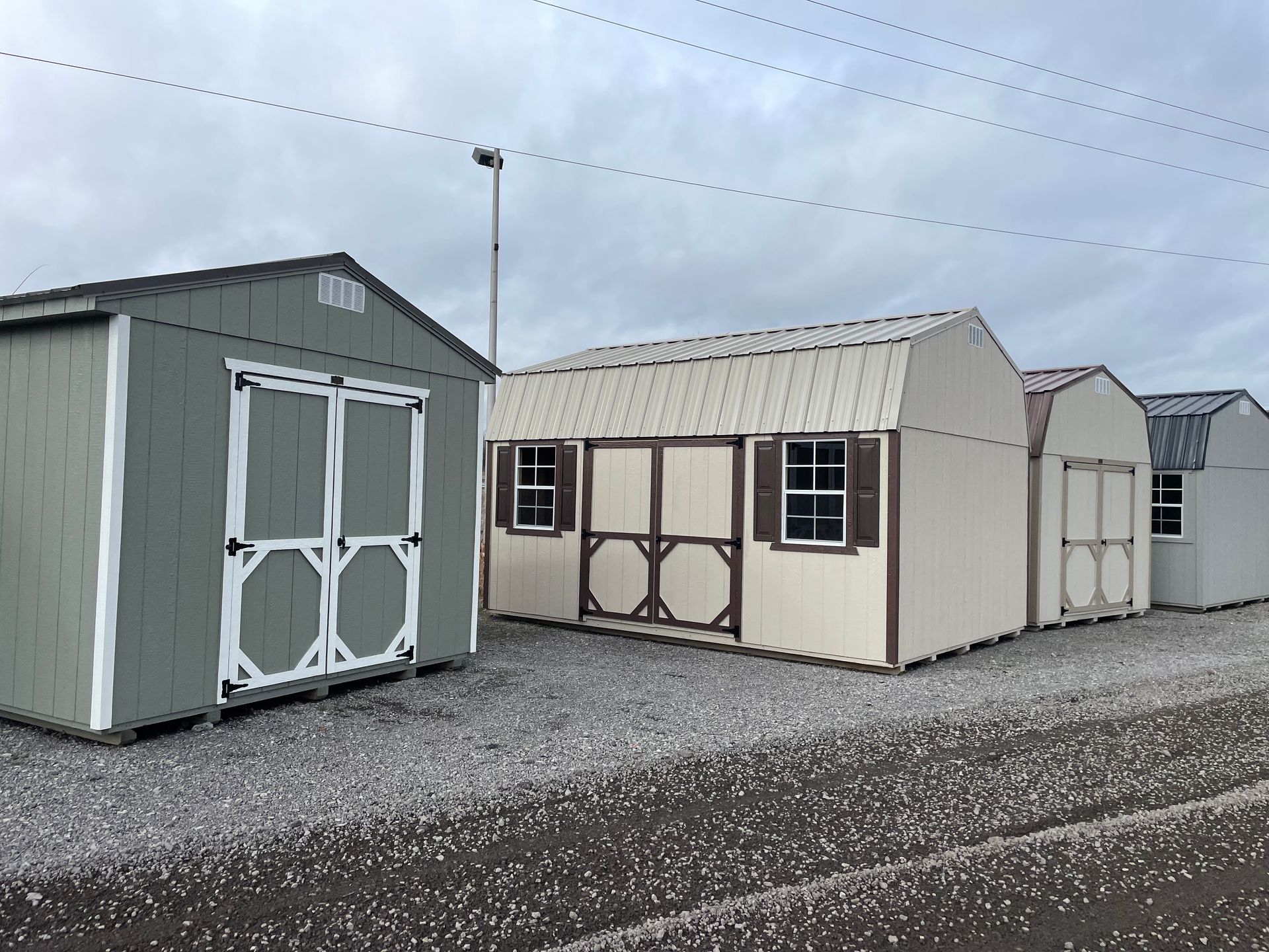 A row of four small outdoor storage sheds in varying colors sitting on a gravel lot under an overcast sky.