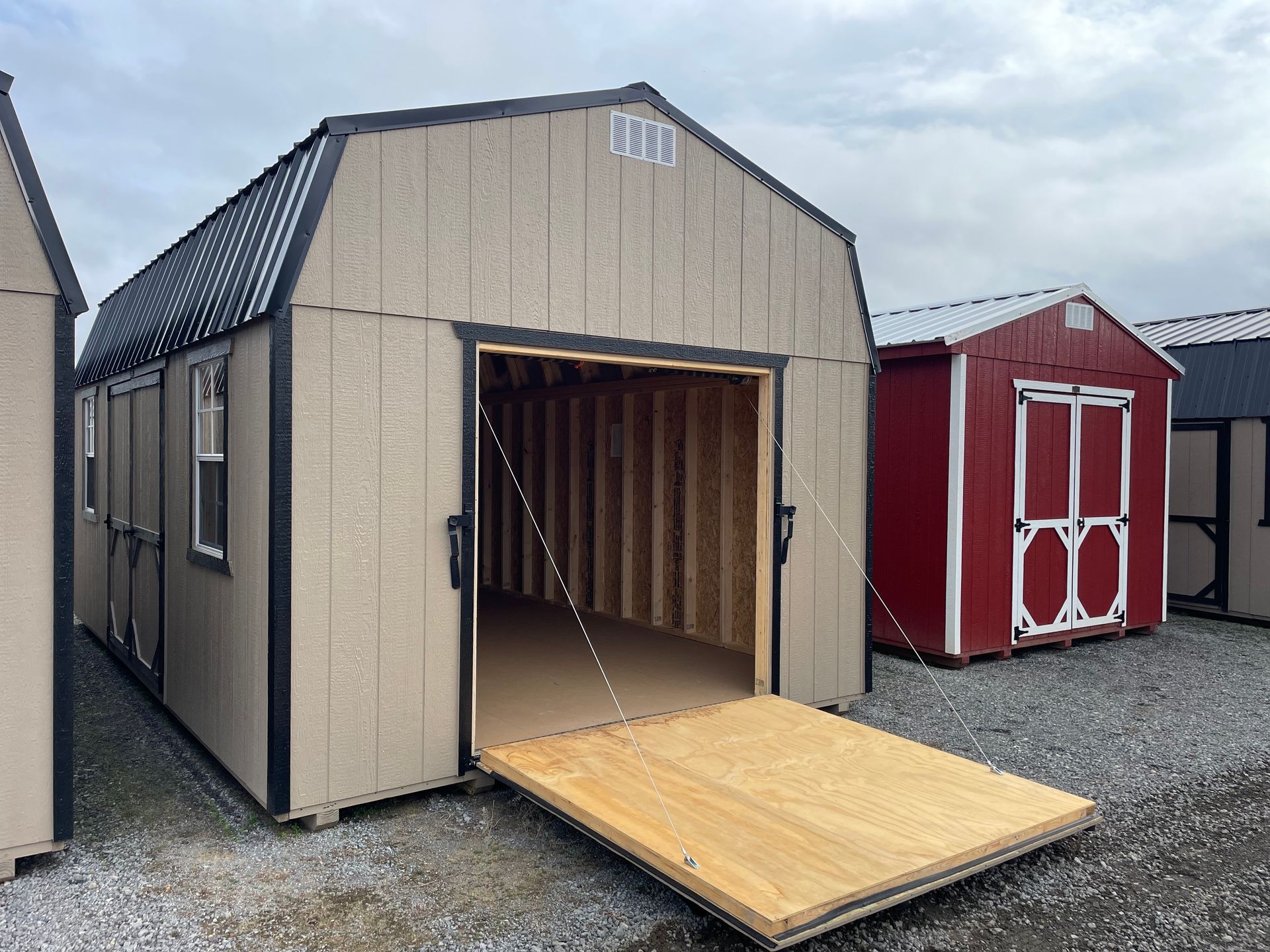 A tan, gambrel-roof shed with an open door and wooden ramp sits on a gravel lot next to a smaller red shed.