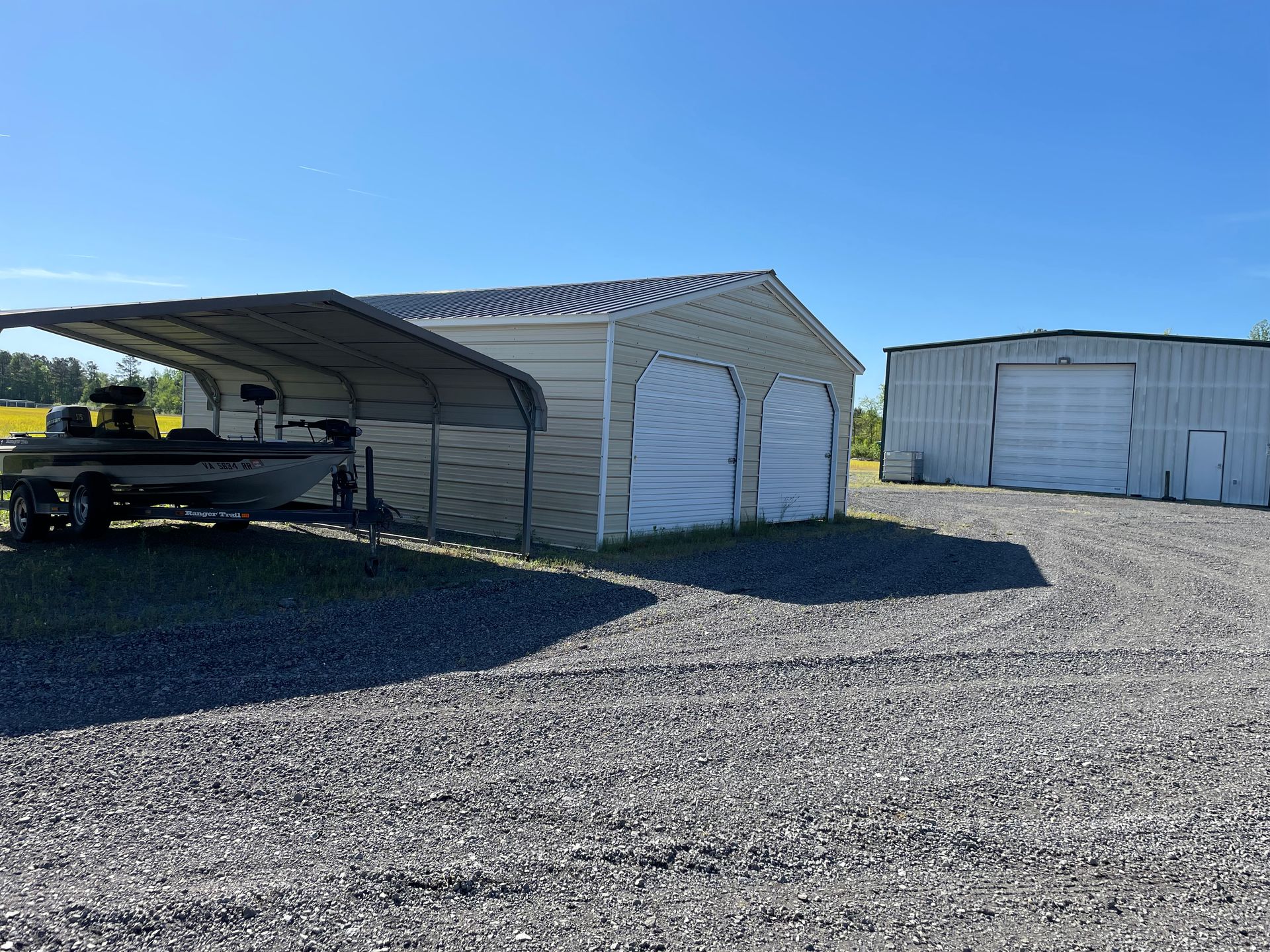 A motorboat sits under a metal carport next to a tan storage building, with a larger white metal building in the distance.