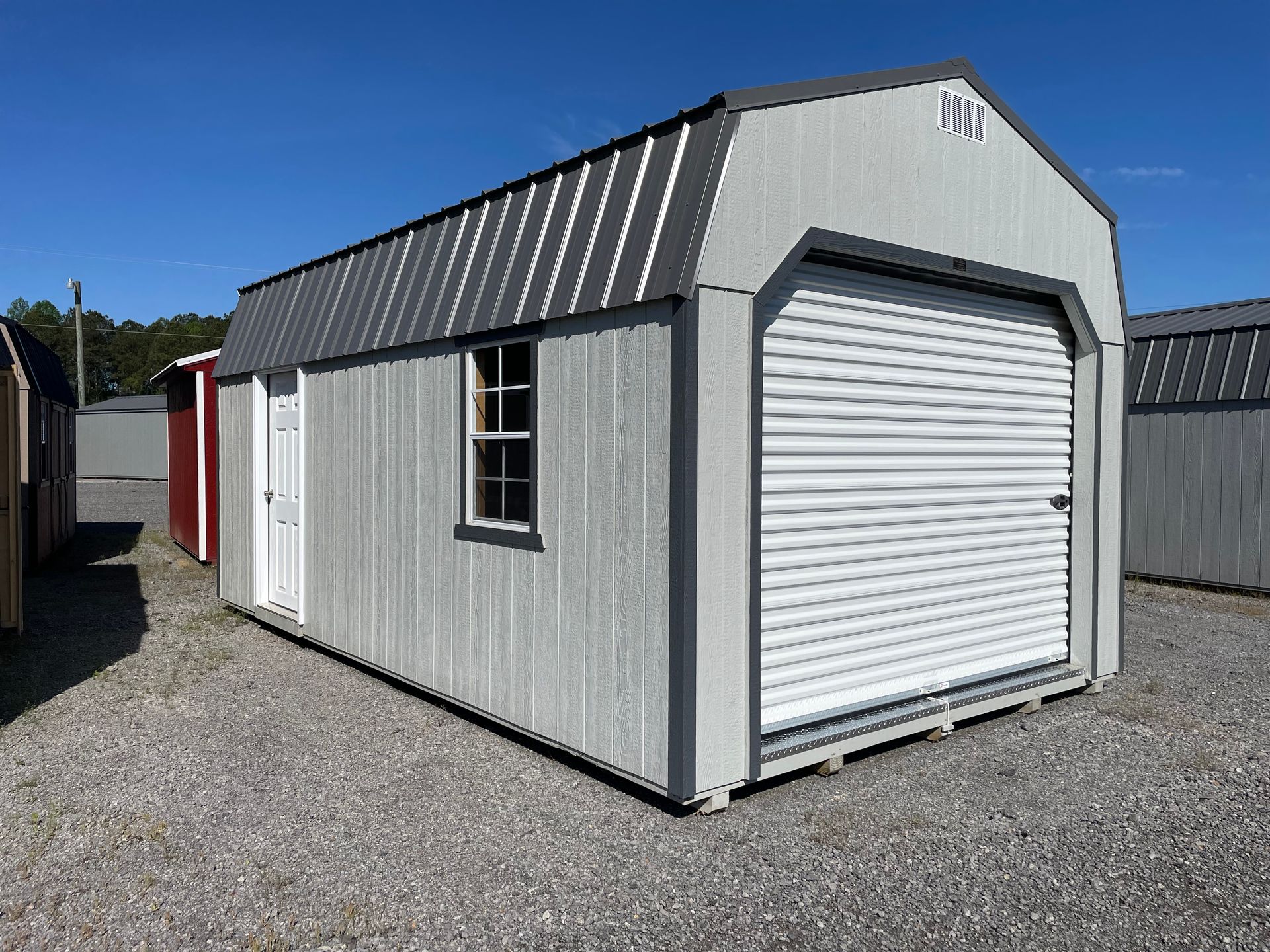 A light gray shed with a dark gray metal roof, featuring a white roll-up garage door and a side entry door.