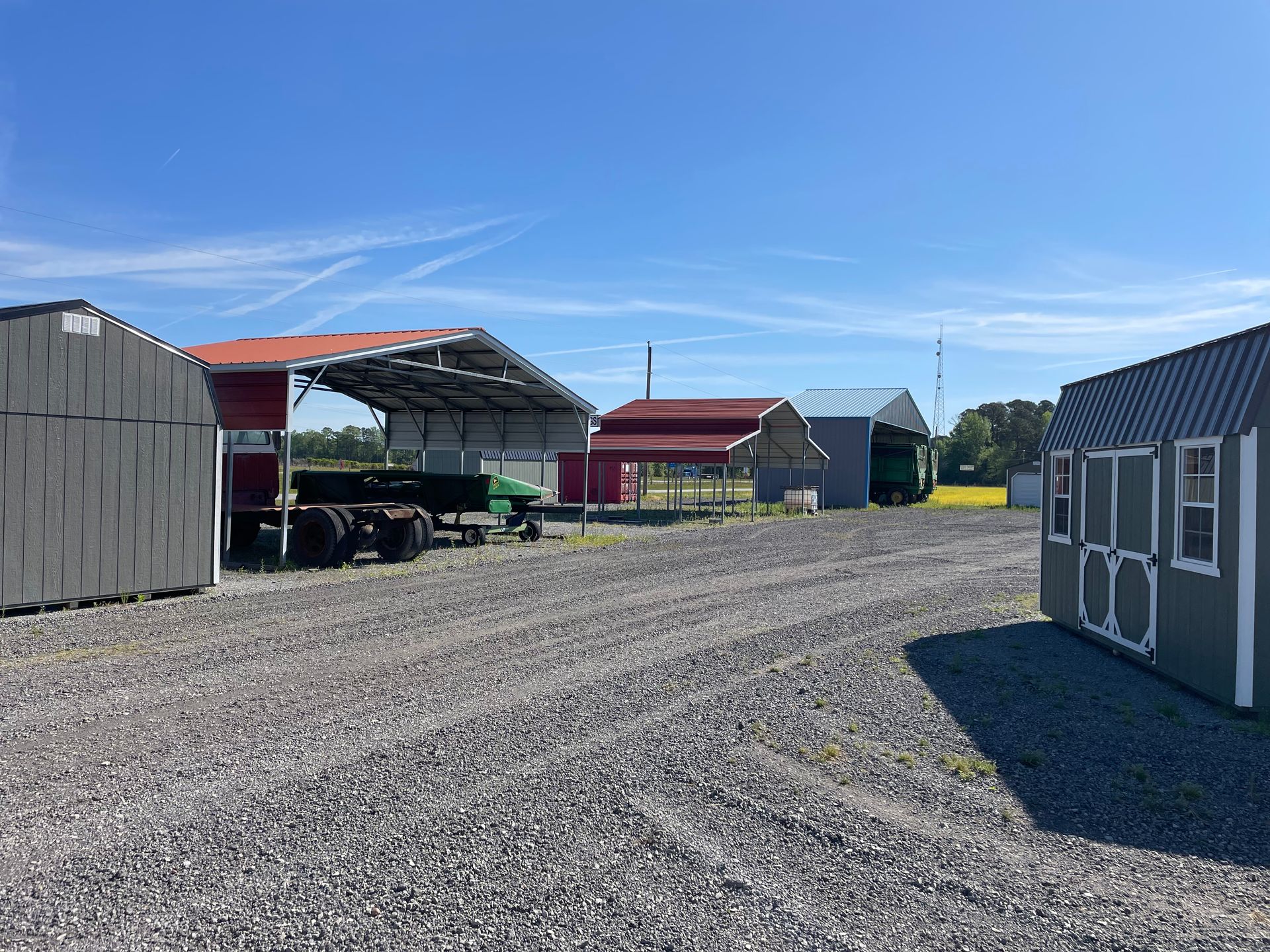 A gravel lot with several sheds, a metal carport sheltering a trailer, and structures under a clear blue sky.