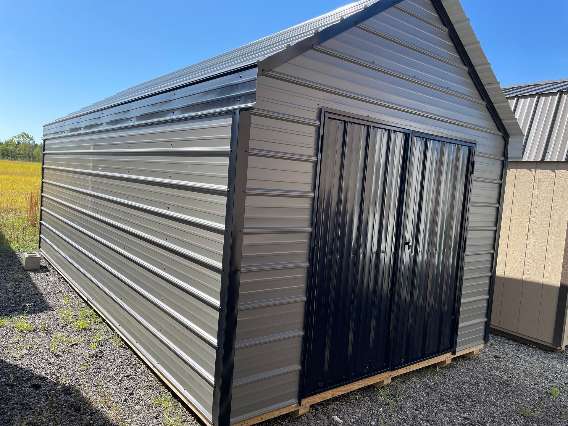 A metal storage shed with grey corrugated walls, black trim, and a black double door, sitting on a gravel lot.