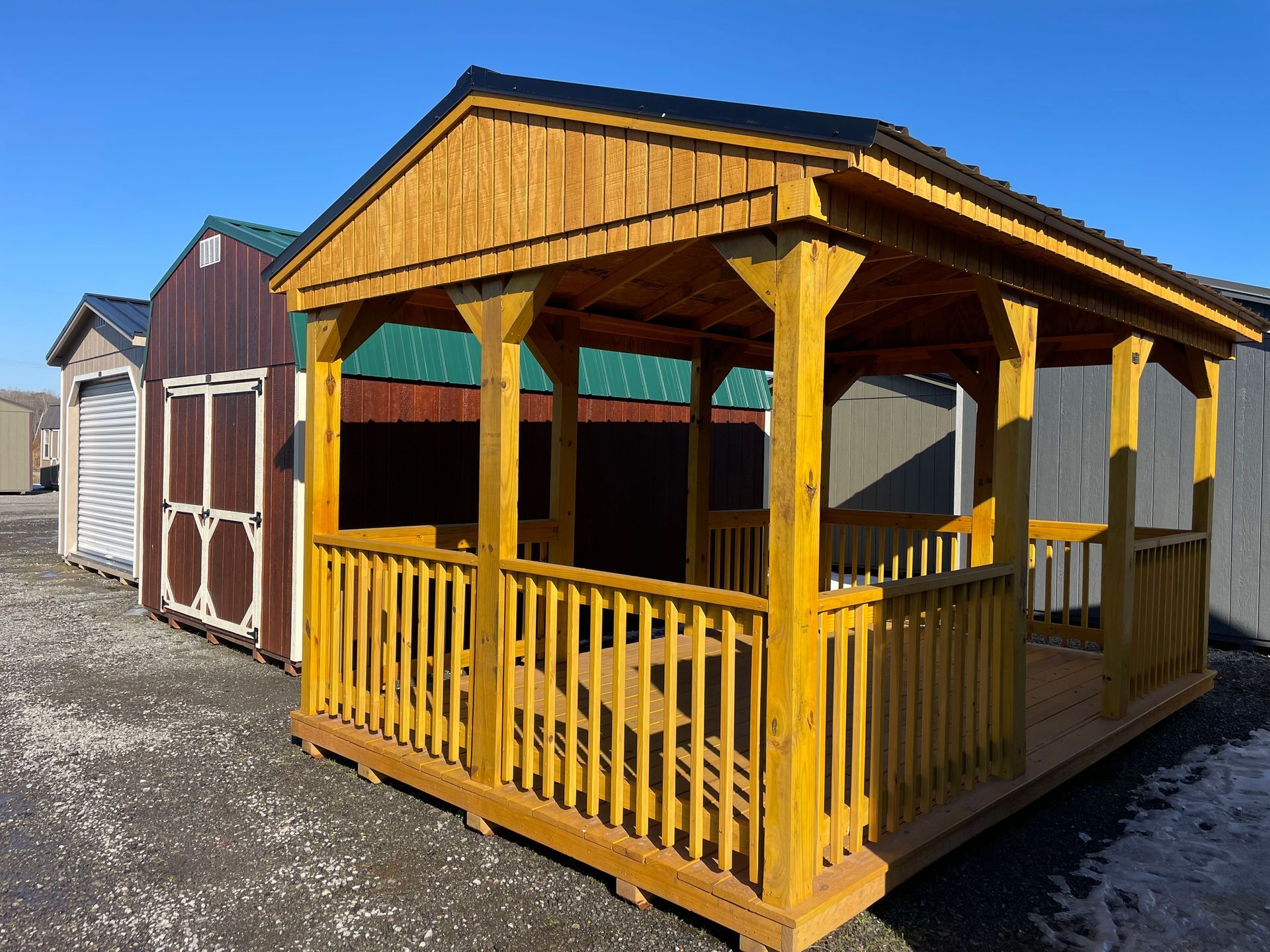 A golden-brown wooden gazebo with a railing sits in a gravel lot next to two storage sheds under a clear blue sky.