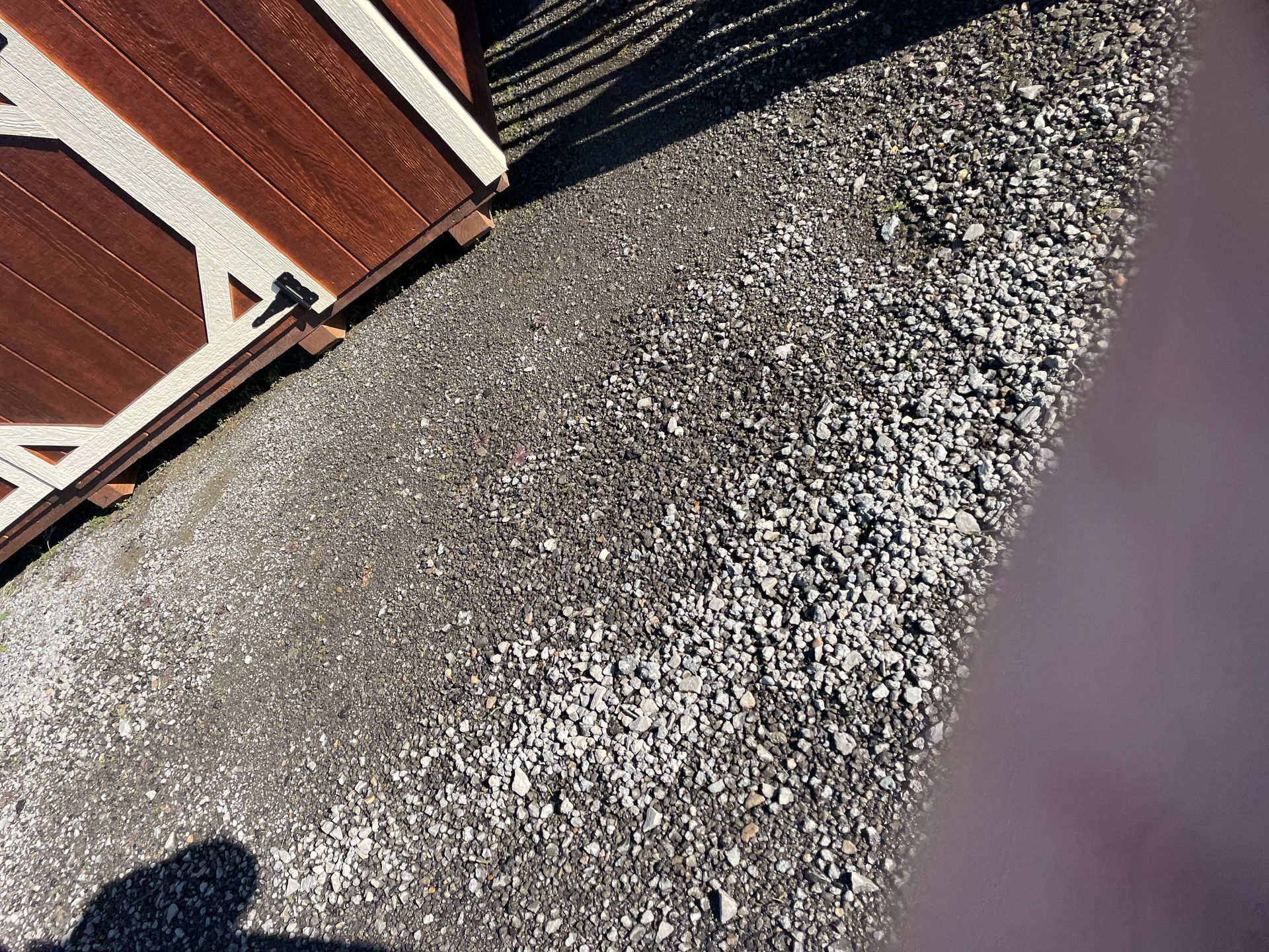 A corner of a brown and white shed sits on a gravel driveway, with a dark shadow cast on the ground.