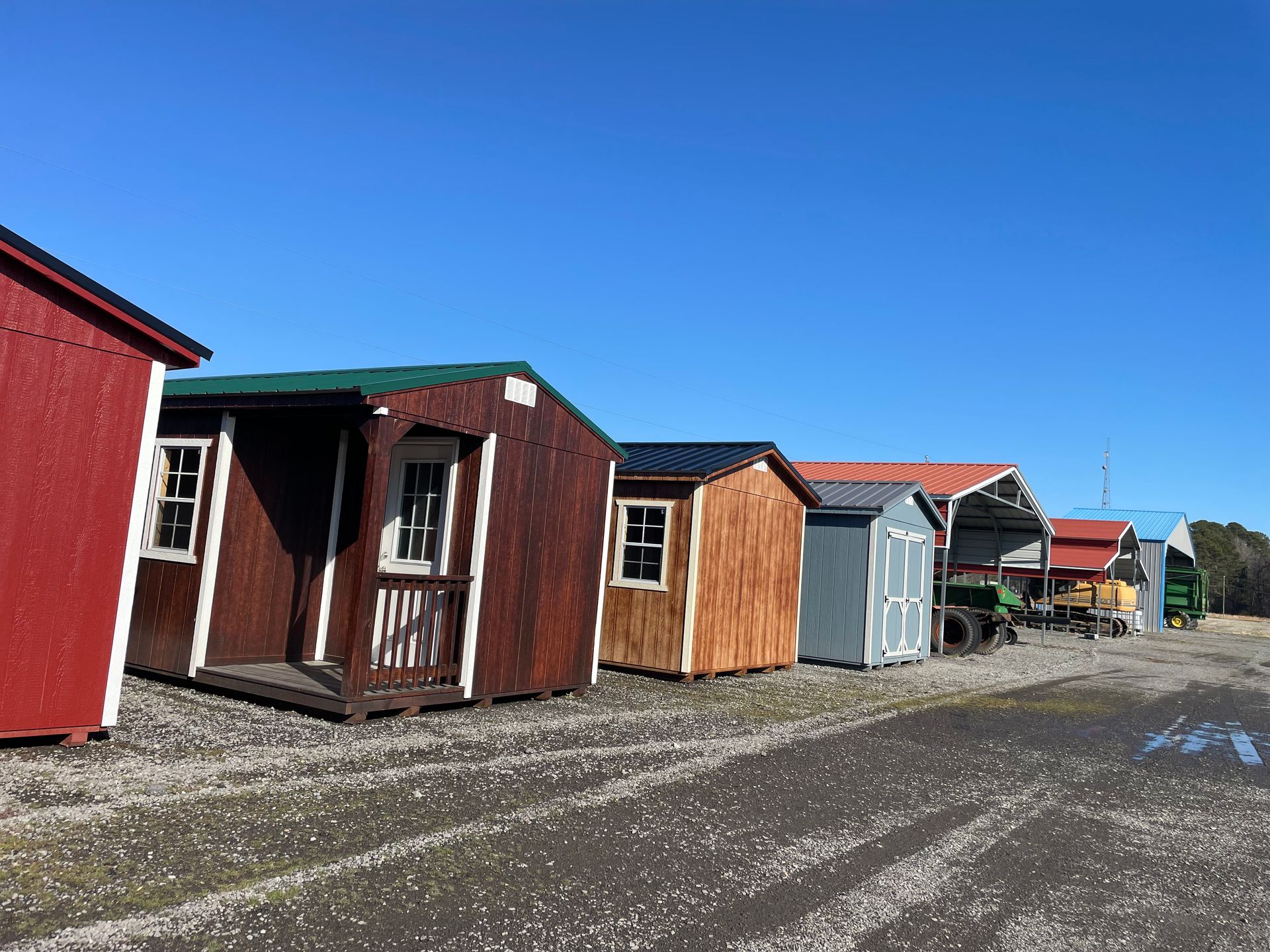 A row of various small sheds and structures for sale on a gravel lot under a clear blue sky.
