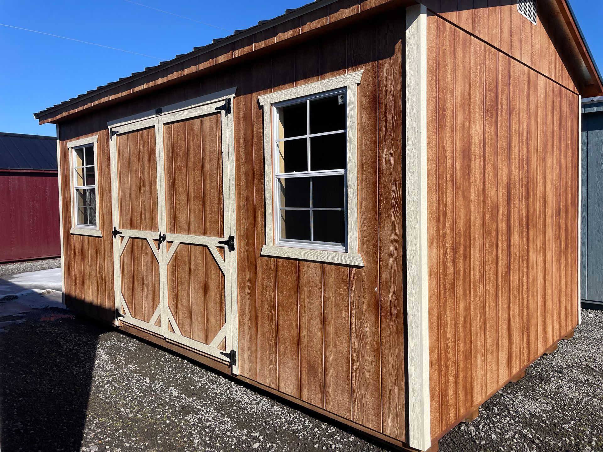 A brown, wooden storage shed with a double door, two windows, and light-colored trim, sitting on a gravel lot.