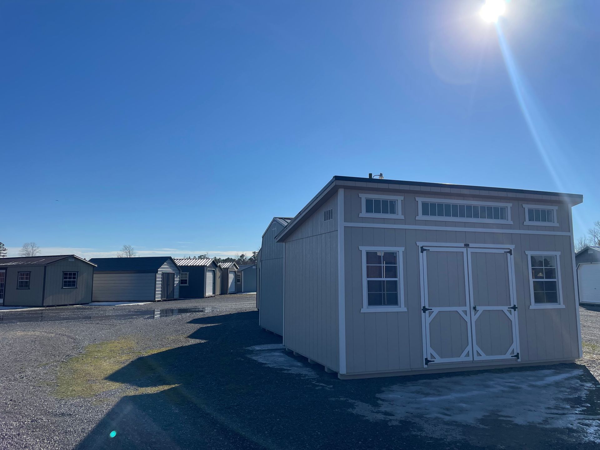 A light-colored storage shed with white trim sits in a gravel lot under a bright blue sky, with other sheds in the distance.
