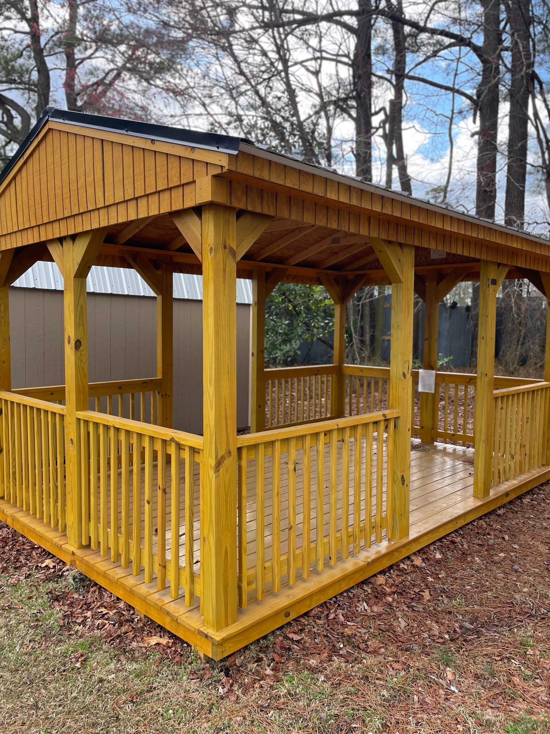 A wooden pavilion with a peaked roof and railings, set outdoors among trees and fallen leaves.