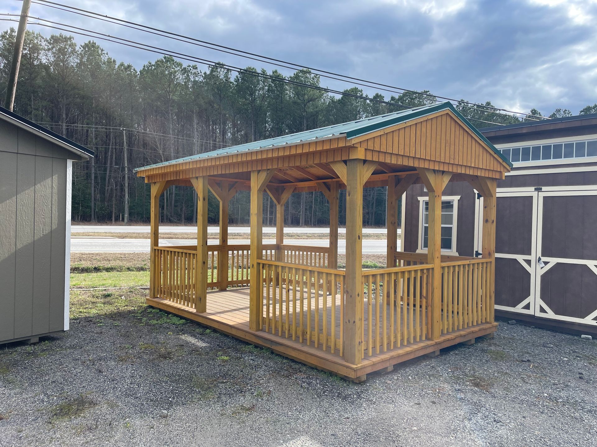 A wooden gazebo with a metal roof and railings, positioned on a gravel lot next to storage sheds.