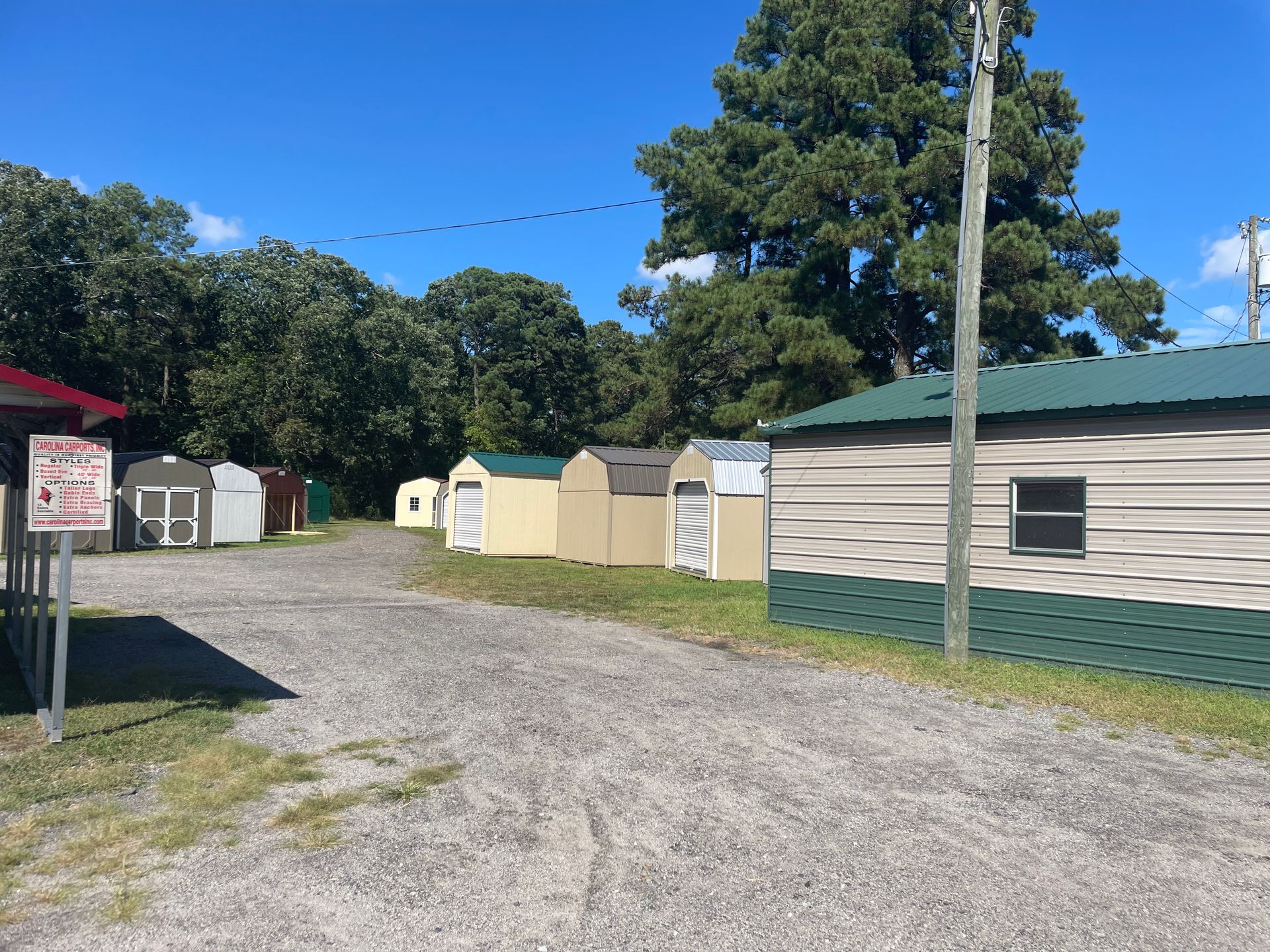 A gravel lot featuring several small, shed-style storage buildings under a clear blue sky, bordered by trees.
