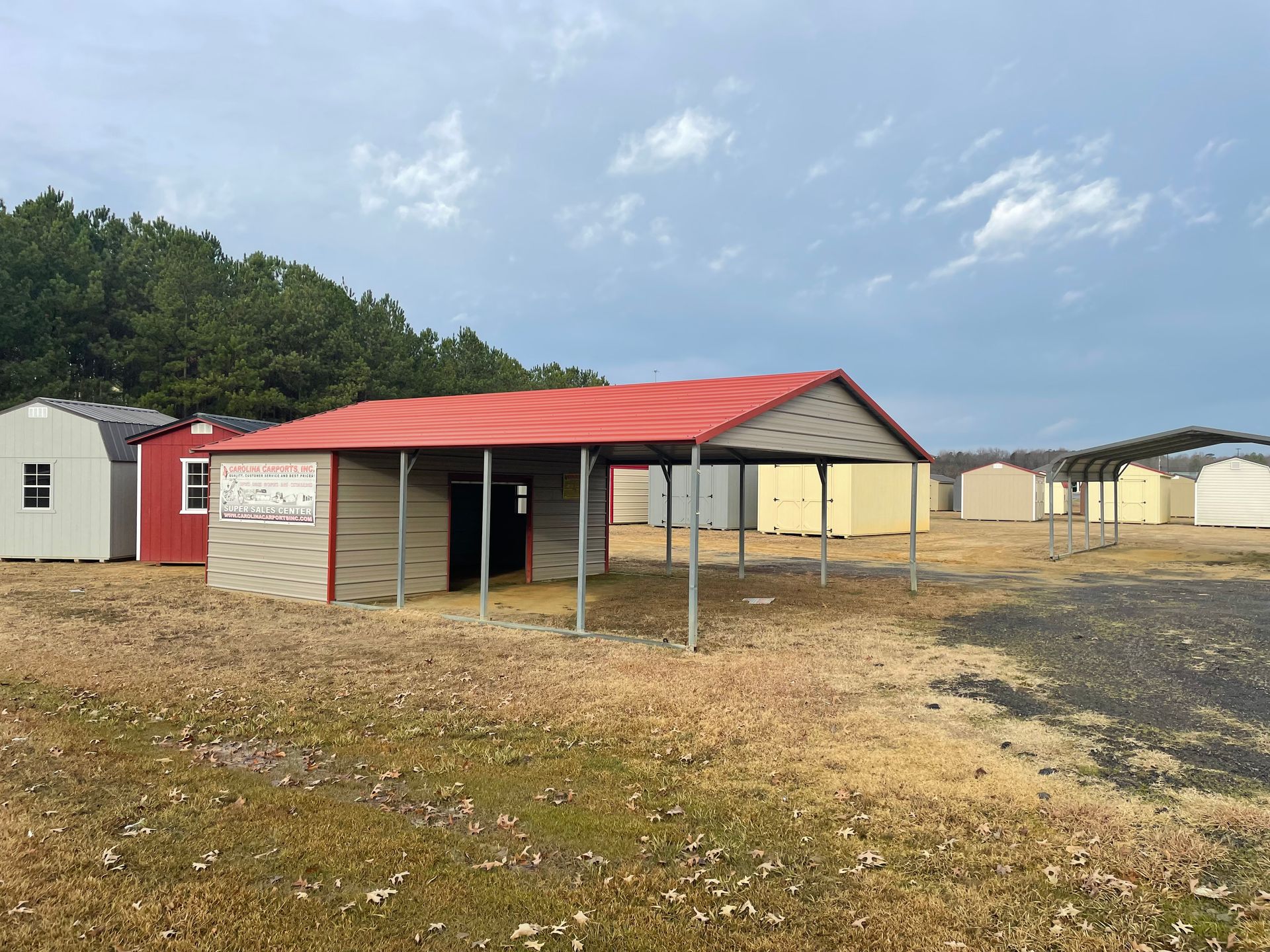 A metal shed with a red roof stands in an open lot alongside several other portable storage buildings on a sunny day.