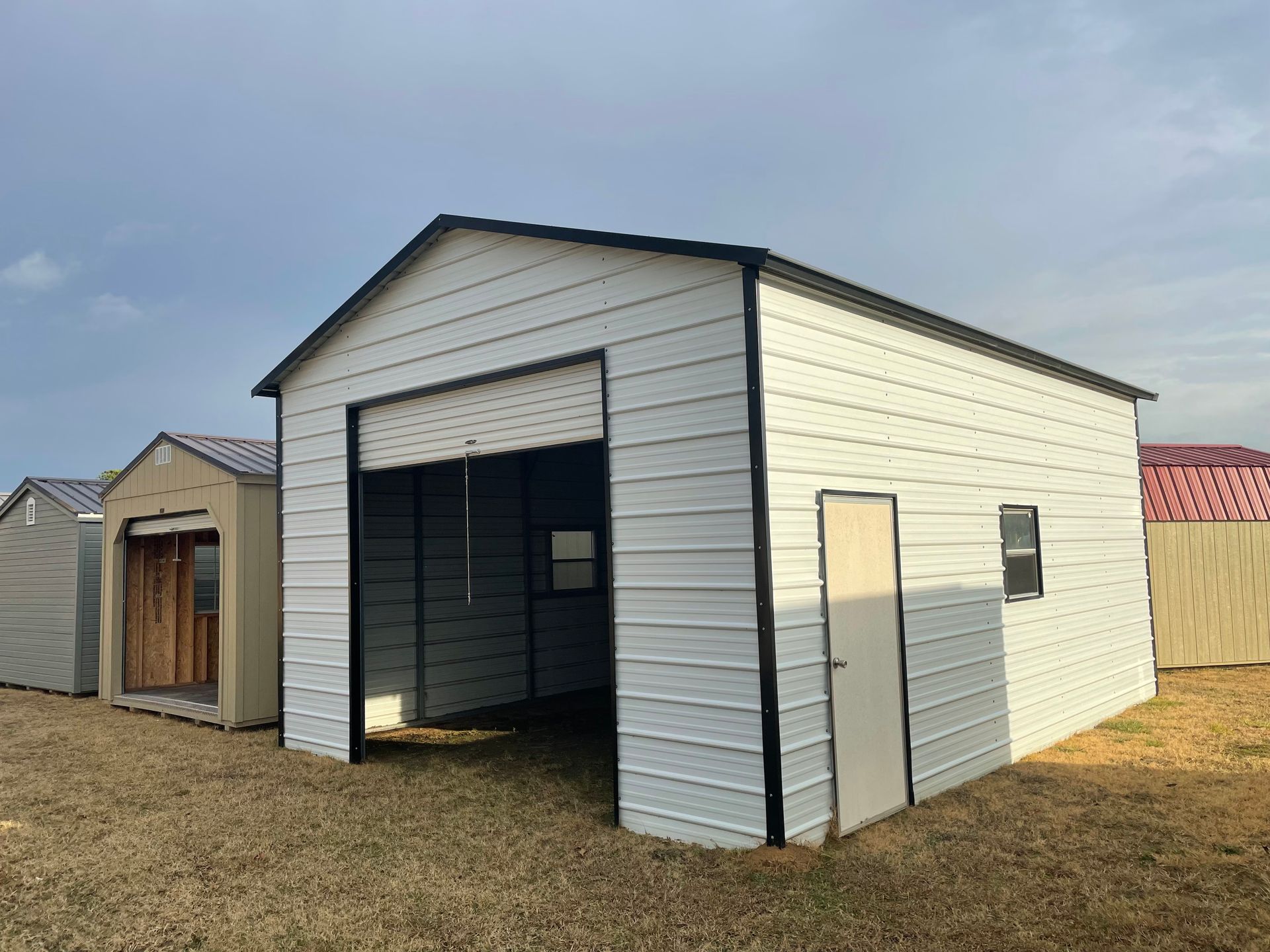 A white metal garage with a roll-up door and a side entry door, standing in a grassy field with other storage sheds.