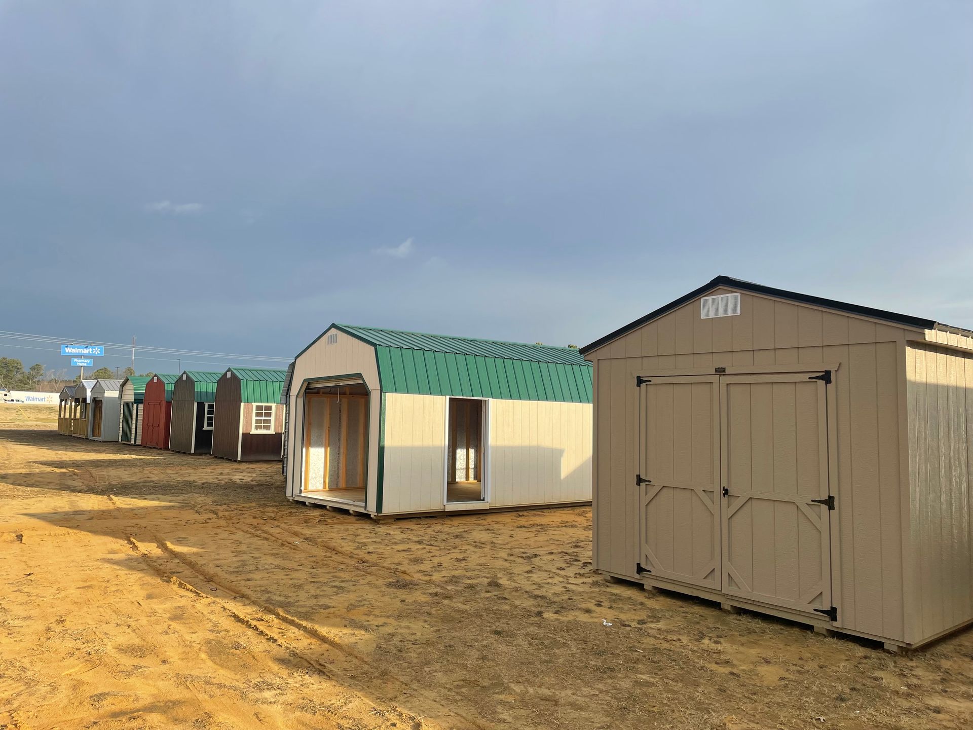A row of several pre-fabricated storage sheds in a sandy lot under a cloudy sky.