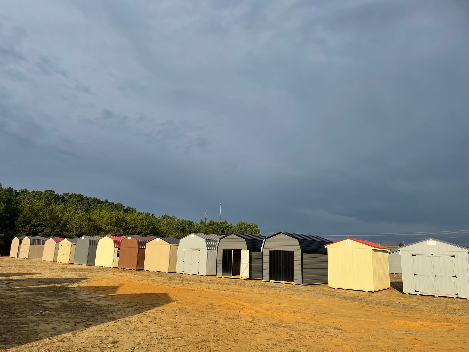 A row of diverse storage sheds in various colors, including white, tan, and gray, arranged on a dirt field under a cloudy sky.