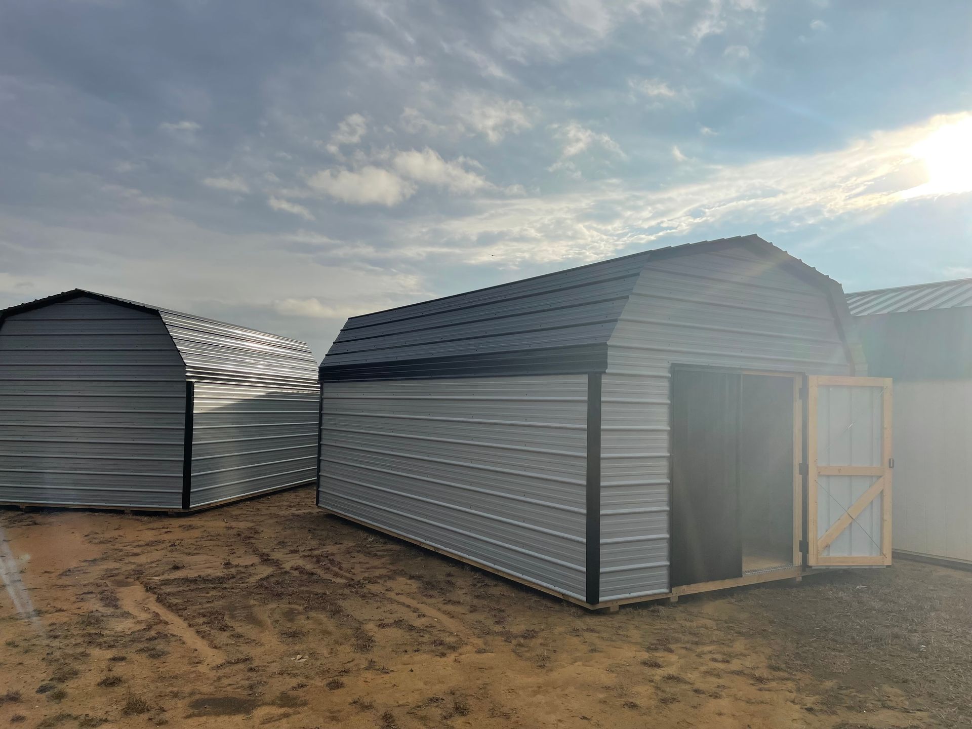 Two gray, metal, barn-style storage sheds sit on a dirt lot under a cloudy, bright sky.