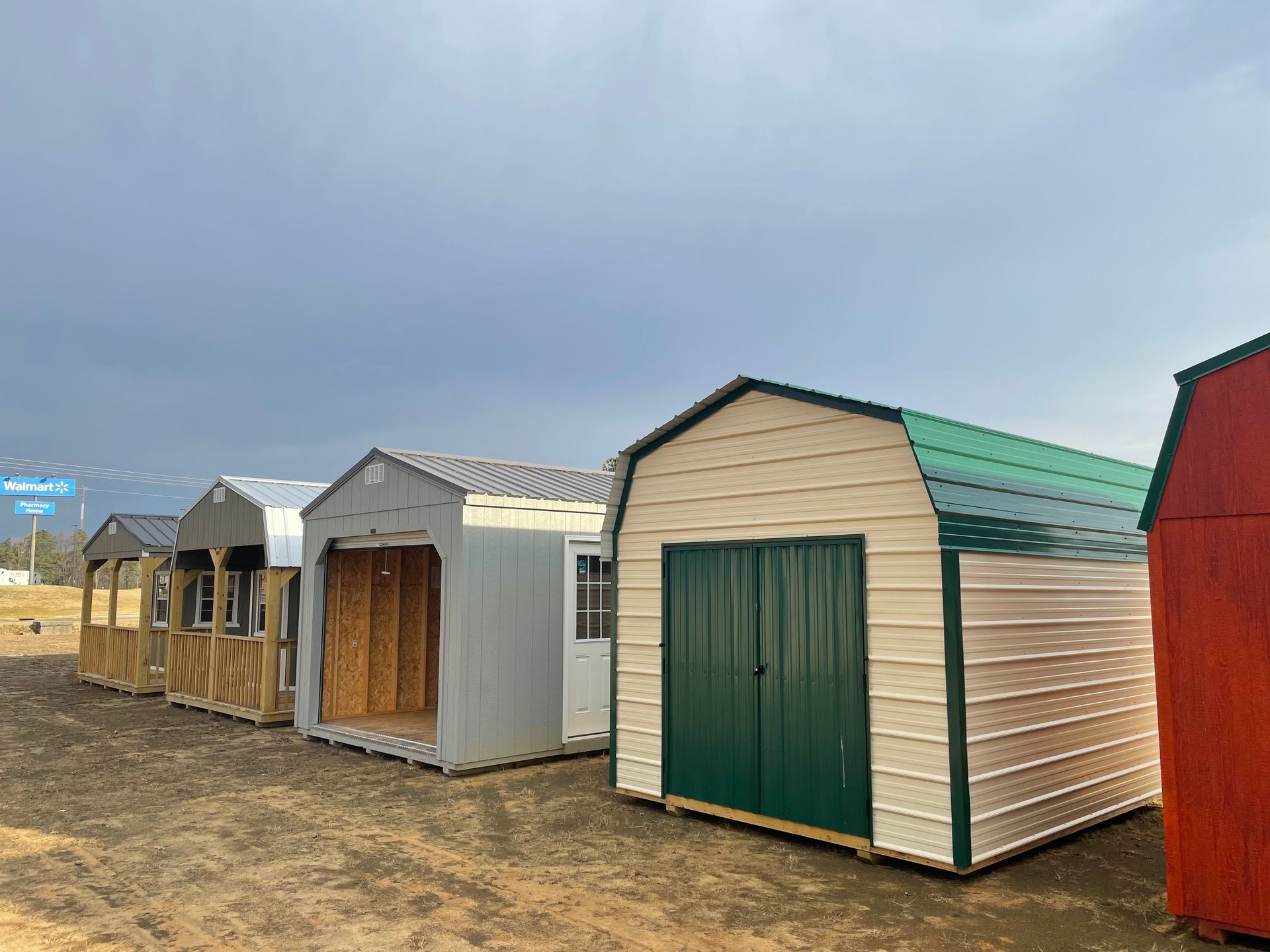 A row of various prefabricated storage sheds in a dirt lot under a cloudy, overcast sky.