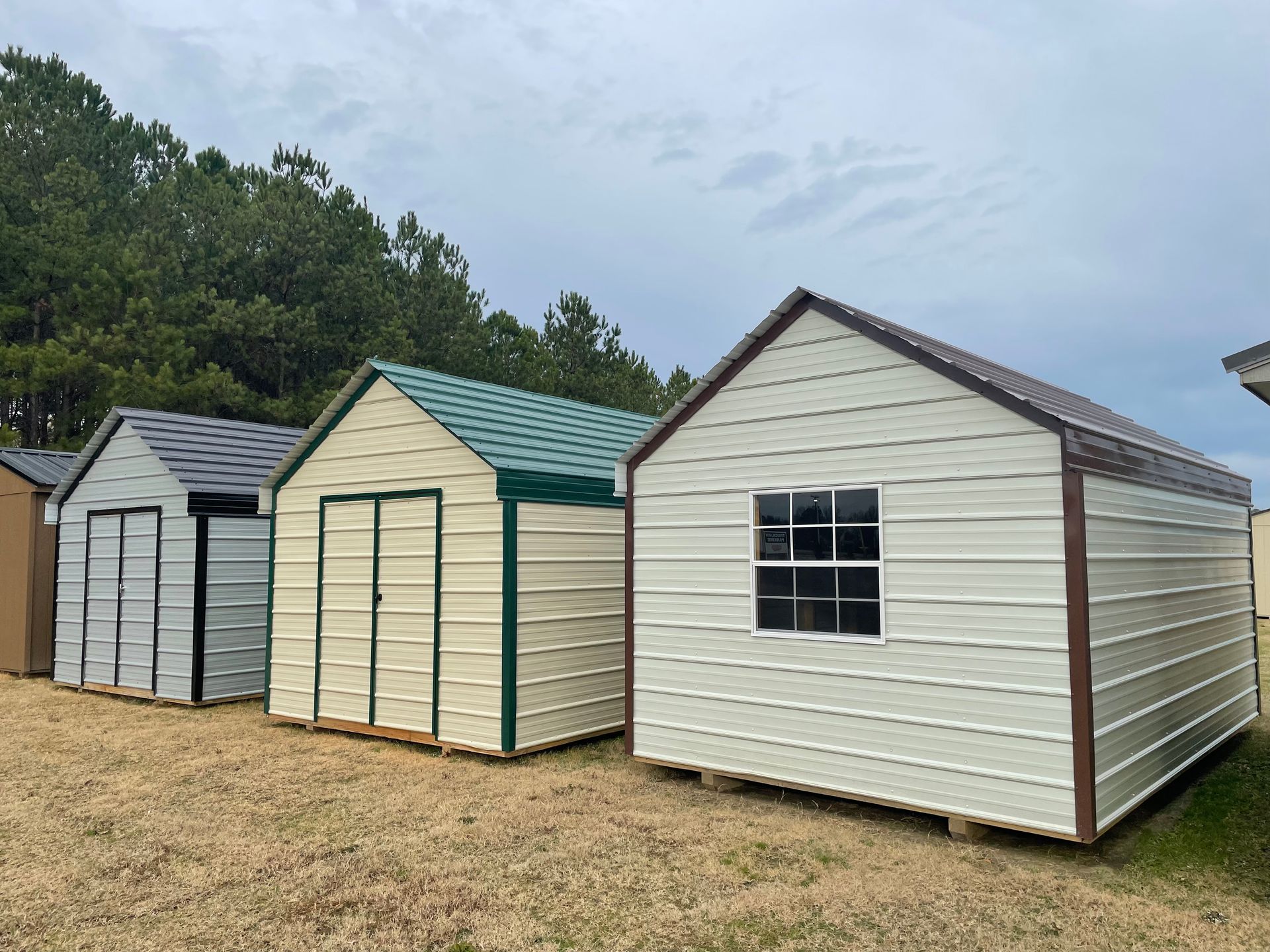 A row of three metal storage sheds in various colors, standing on a gravel lot in front of trees under a cloudy sky.