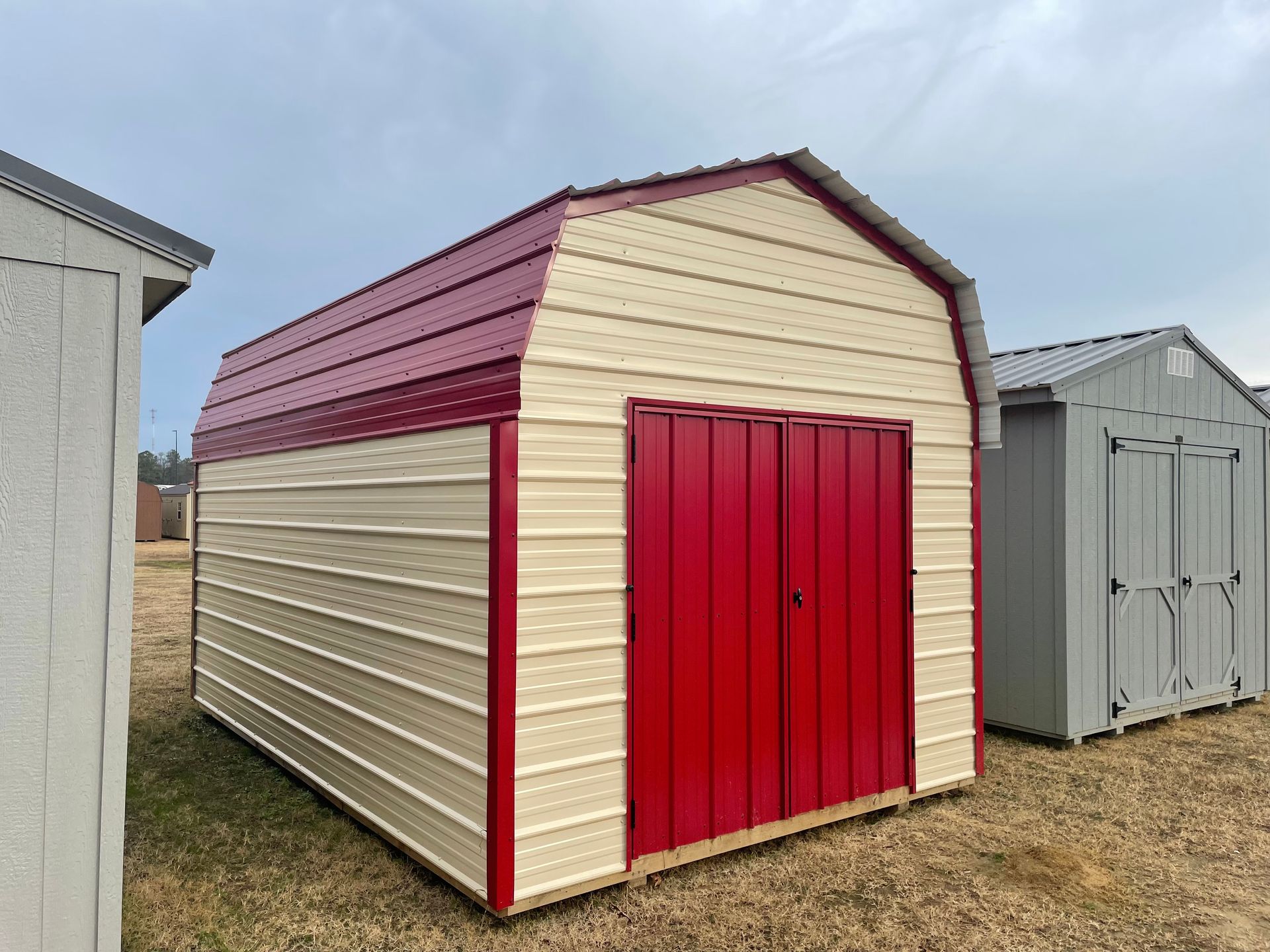A cream-colored metal barn-style storage shed with red doors and a matching red roof, standing in an outdoor lot.