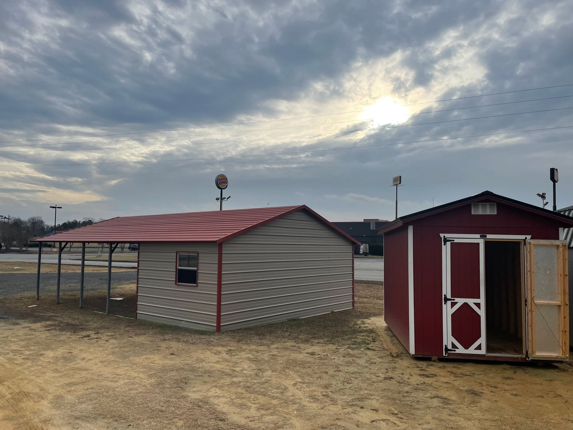 Two outdoor storage sheds, one with a red metal roof and side carport and one smaller red shed, set under a cloudy sky.