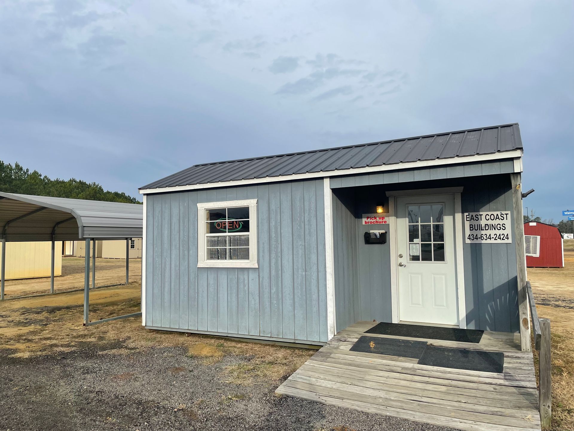 A light blue portable building with a white door, a window, and a dark metal roof, set on a gravel lot under a cloudy sky.