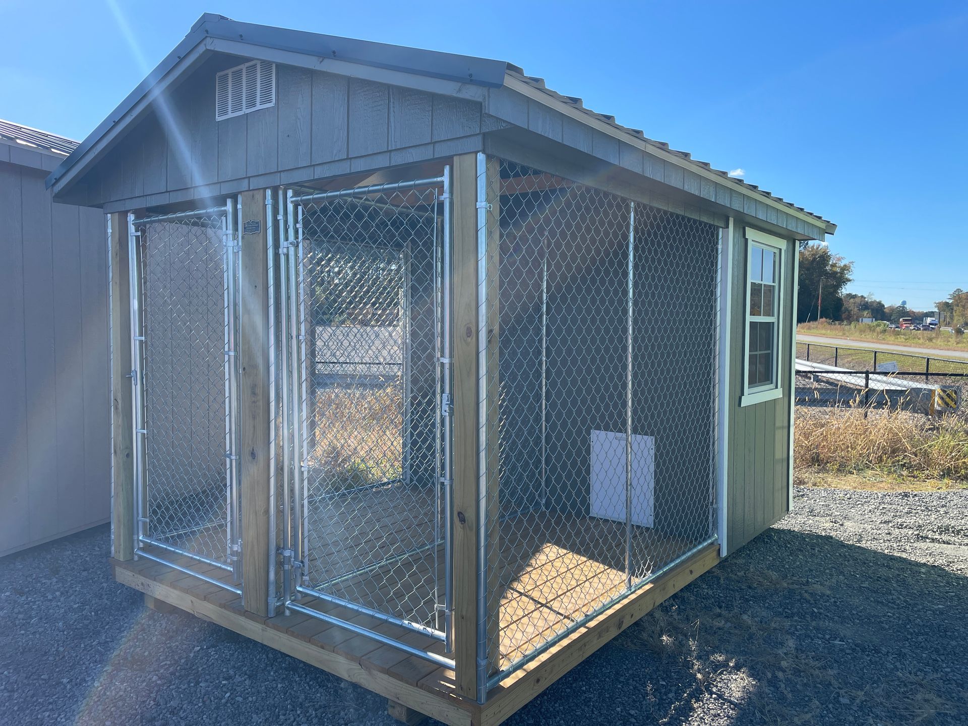 A wooden outdoor kennel with a chain-link fenced front area, metal roof, and window, set on a gravel surface.