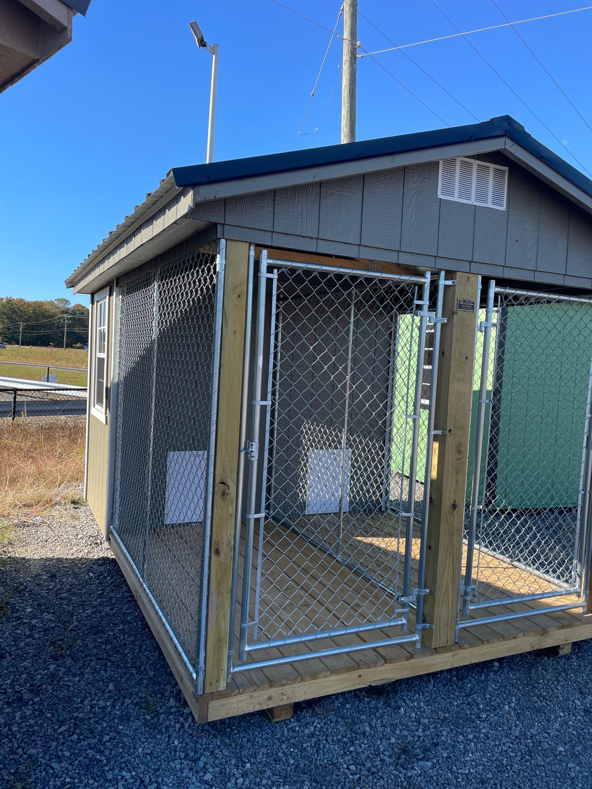 A gray wooden outdoor dog kennel with chain-link fencing, doors, and a wood floor on a gravel surface under a blue sky.
